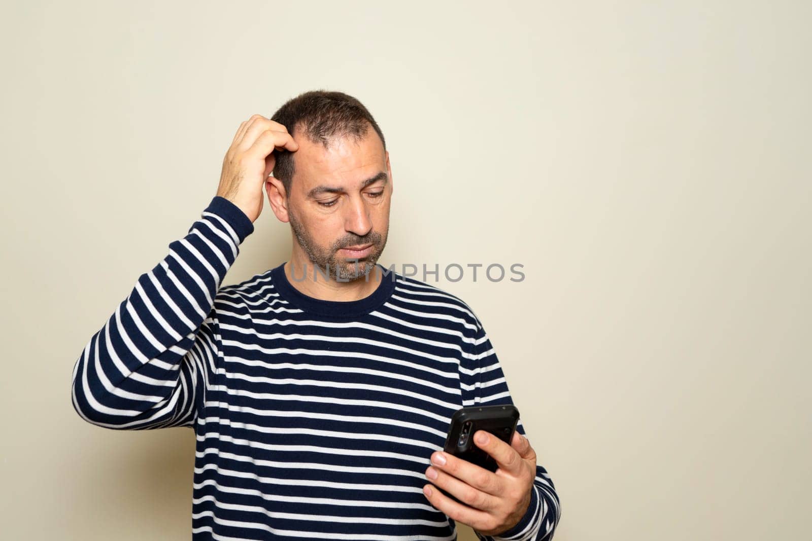 Bearded Hispanic man in his 40s wearing a striped sweater scratching his head while searching for the solution to a problem on his smartphone isolated over beige background. by Barriolo82