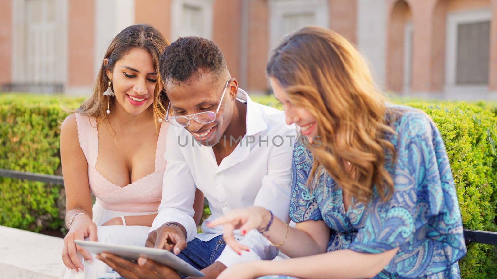 Smiling friends sitting on the street while using a tablet by ivanmoreno