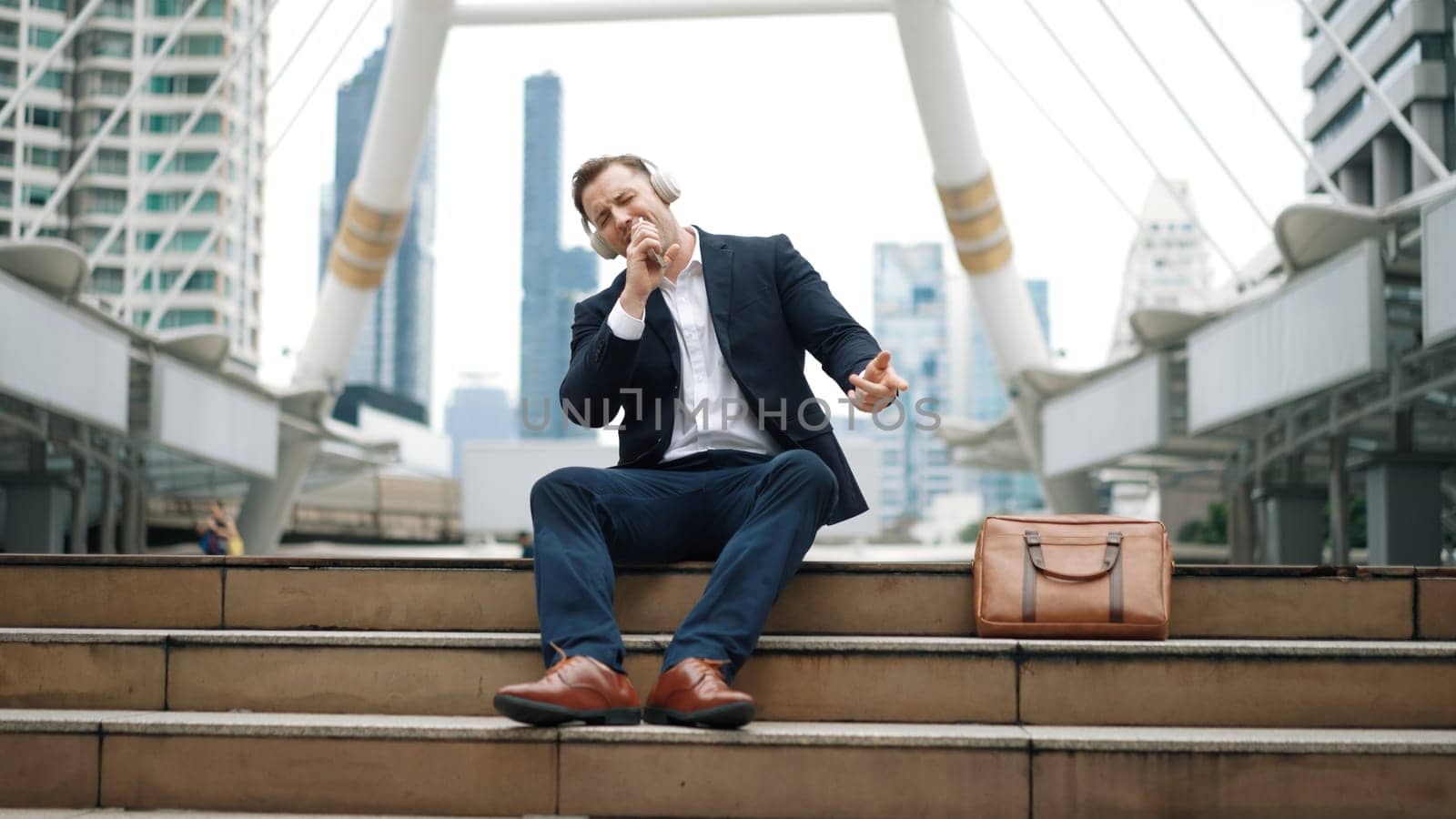 Professional business man listening headphone and using phone record voice while sitting at stair in urban city. Manager using headset listening relaxed song and moving along in lively mood. Urbane.