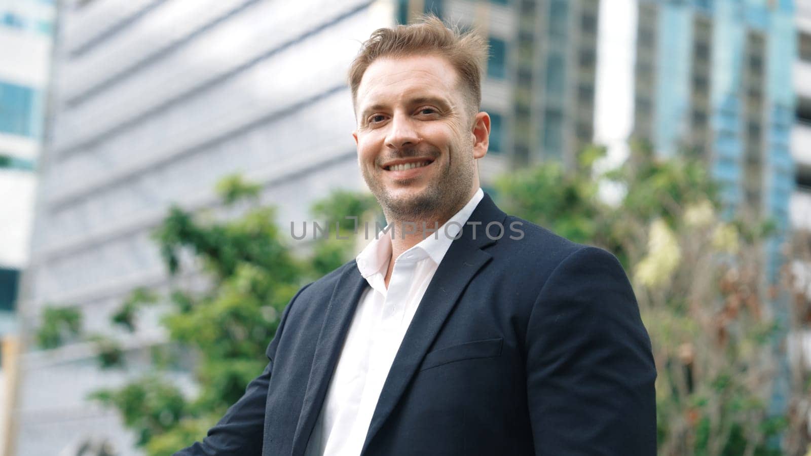 Skilled caucasian business man looking at camera while standing at rooftop. Professional investor smiling with confident while wearing formal suit at modern building in green city. Lifestyle. Urbane