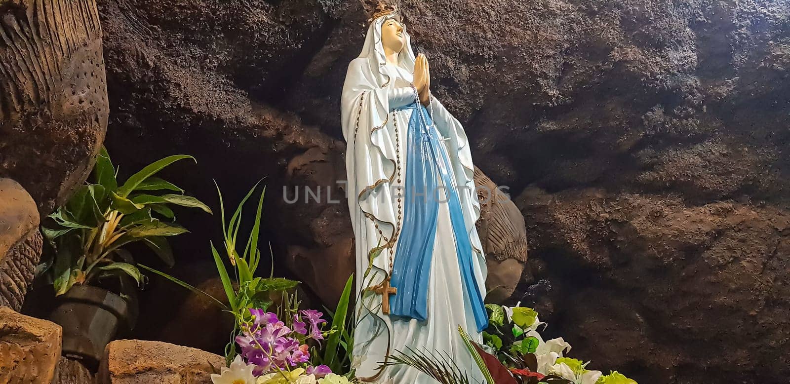 Statue of Holy Virgin Mary in Roman Catholic Church, in the cave of virgin mary, in a rock cave chapel Catholic Church with tropical flowers around in the asian