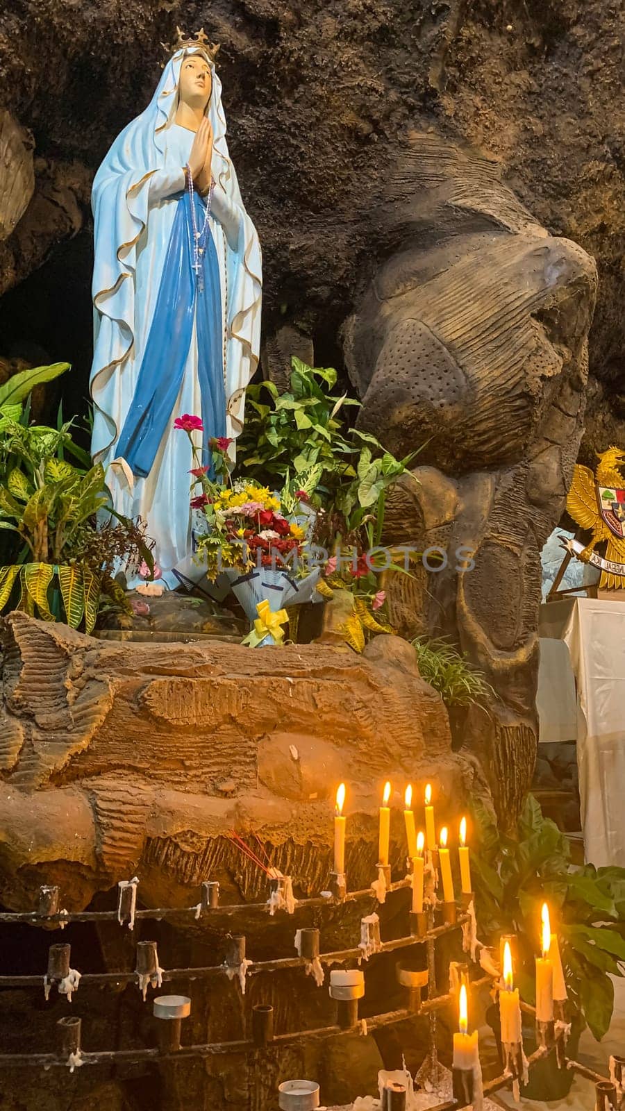 Statue of Holy Virgin Mary in Roman Catholic Church, in the cave of virgin mary, in a rock cave chapel Catholic Church with tropical flowers around in the asian
