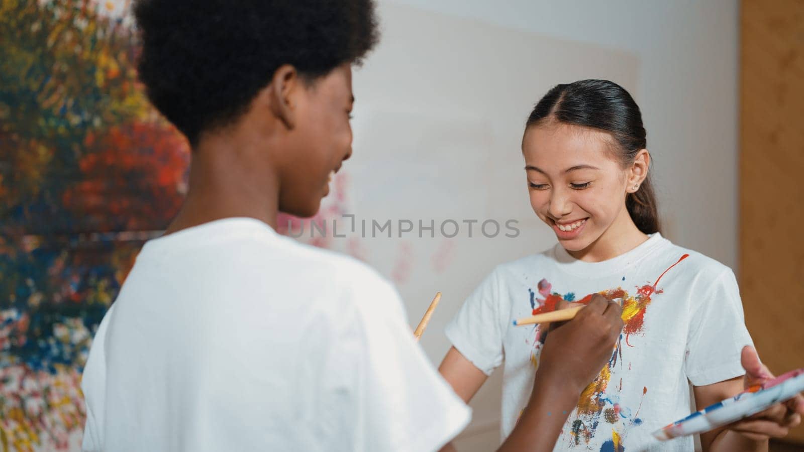 Smiling diverse children using paint brush painted color on each other white shirt shirt at colorful stained wall in art lesson. Represent exchanging experience, learning each other. Edification.