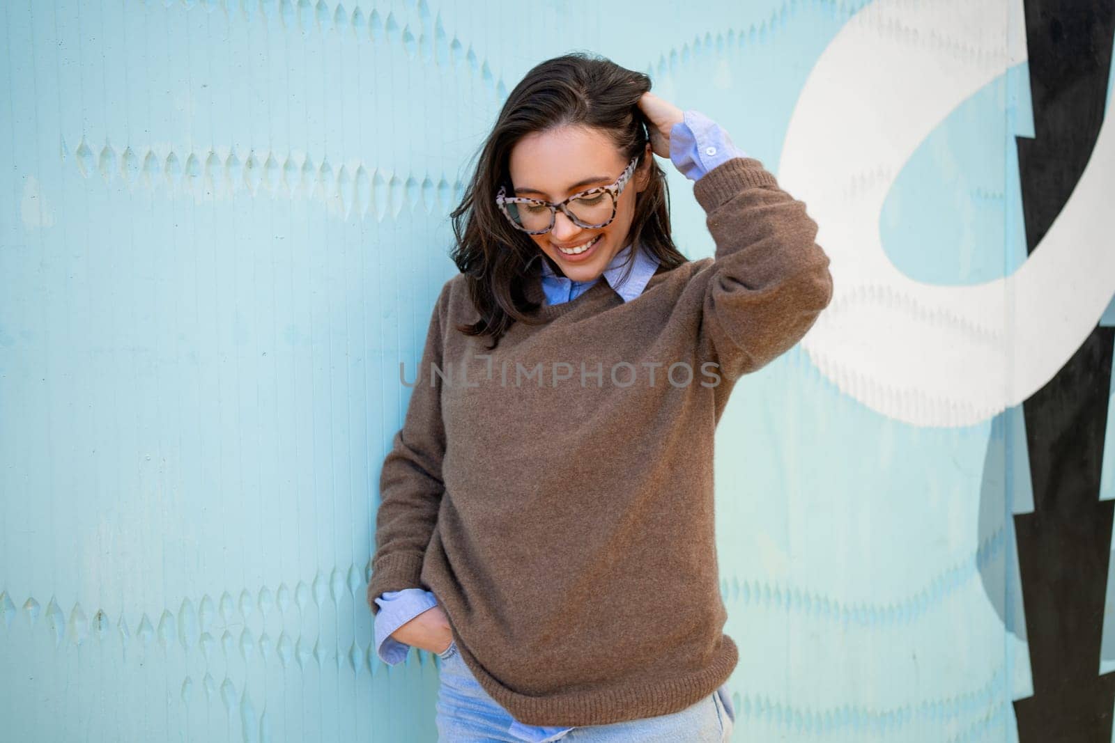 Happy woman in glasses outdoor on blue color background. Positive people concept. Smiling girl looking down, touching hair, hands in pocket, dressed sweater and jeans. Vertical full length