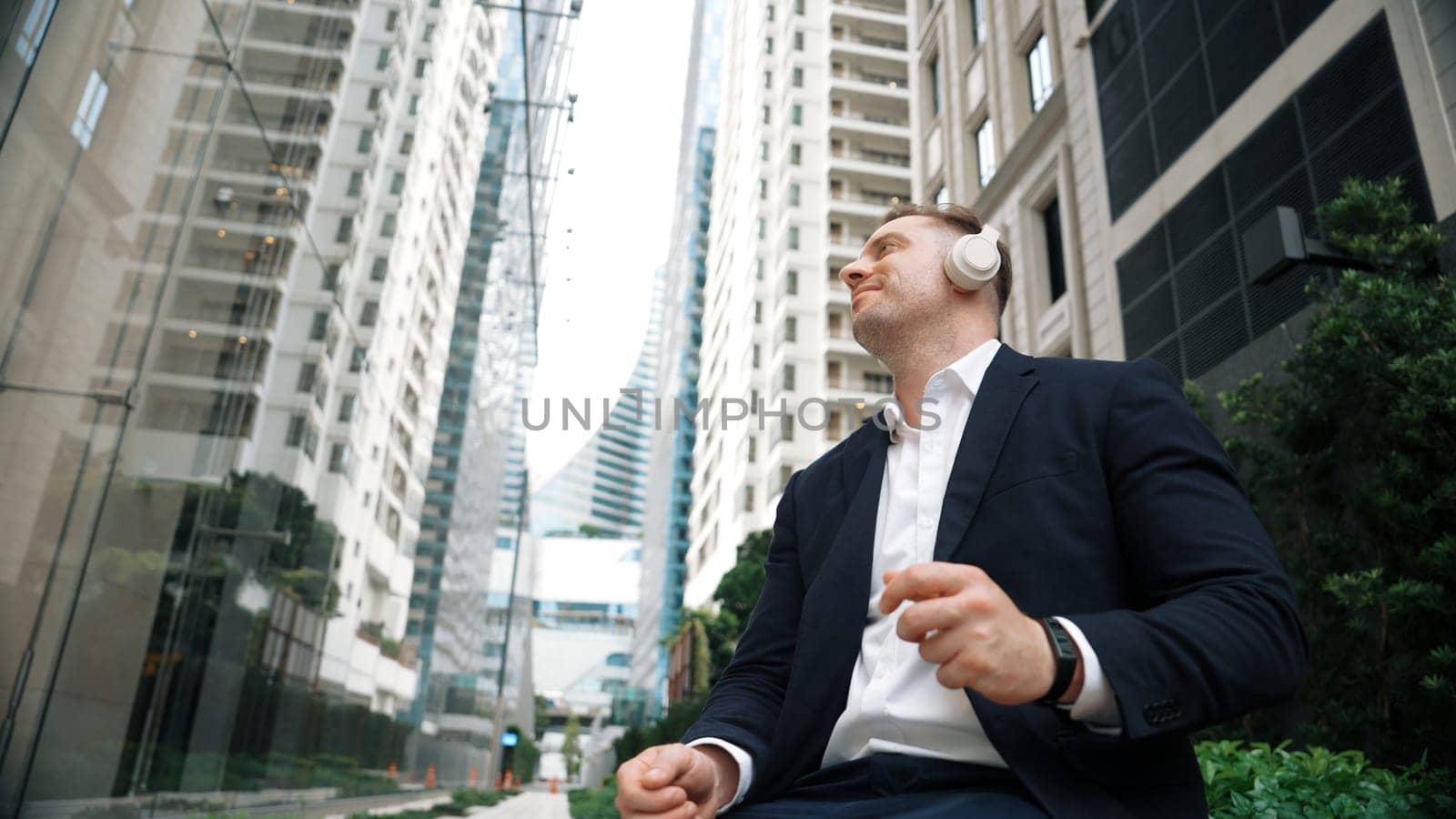 Low angle camera of project manager listening relaxing music while move to song and clap hands. Happy caucasian businessman enjoy listening music while sitting at bench at green urban city. Urbane.