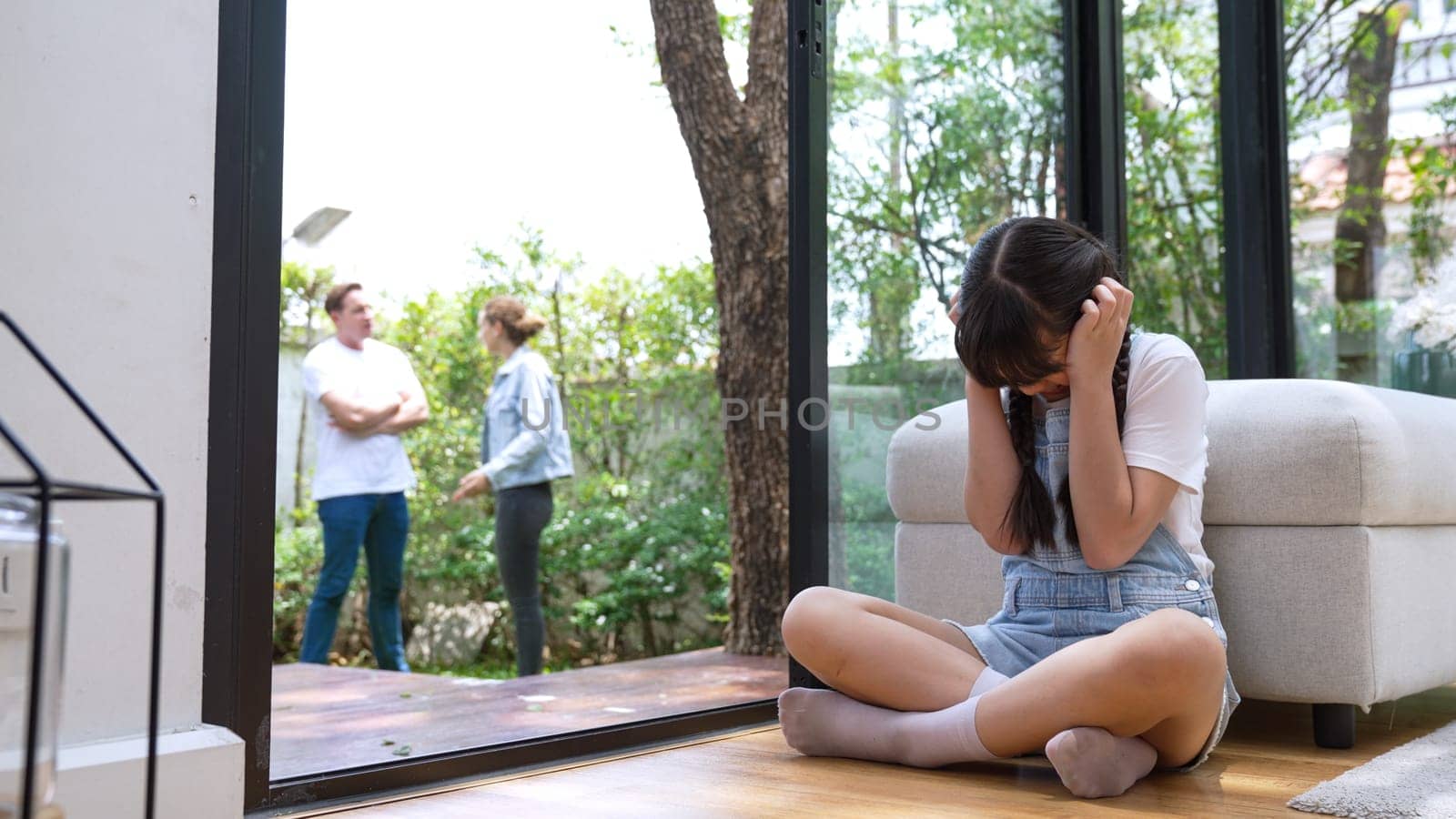 Stressed and unhappy young girl huddle in corner, cover her ears blocking sound of her parent arguing in background. Domestic violence at home and traumatic childhood develop to depression. Synchronos