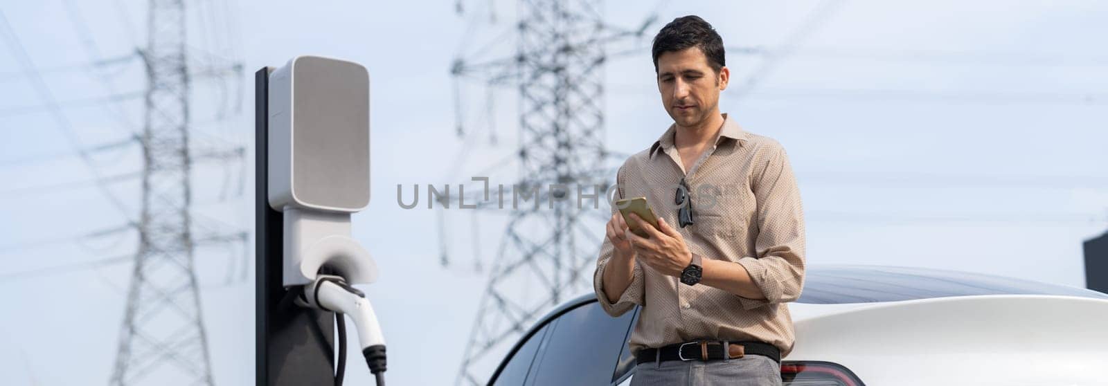 Man pay for electricity with smartphone while recharge EV car battery at charging station connected to electrical [ower grid tower for eco friendly car utilization. Panorama Expedient