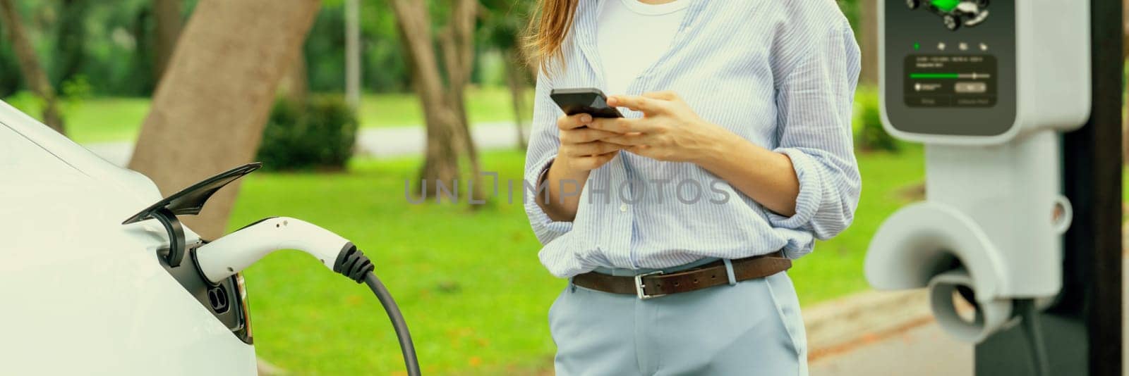 Young woman using smartphone online banking application to pay for electric car battery charging from EV charging station during vacation road trip at national park or summer forest. Panorama Exalt