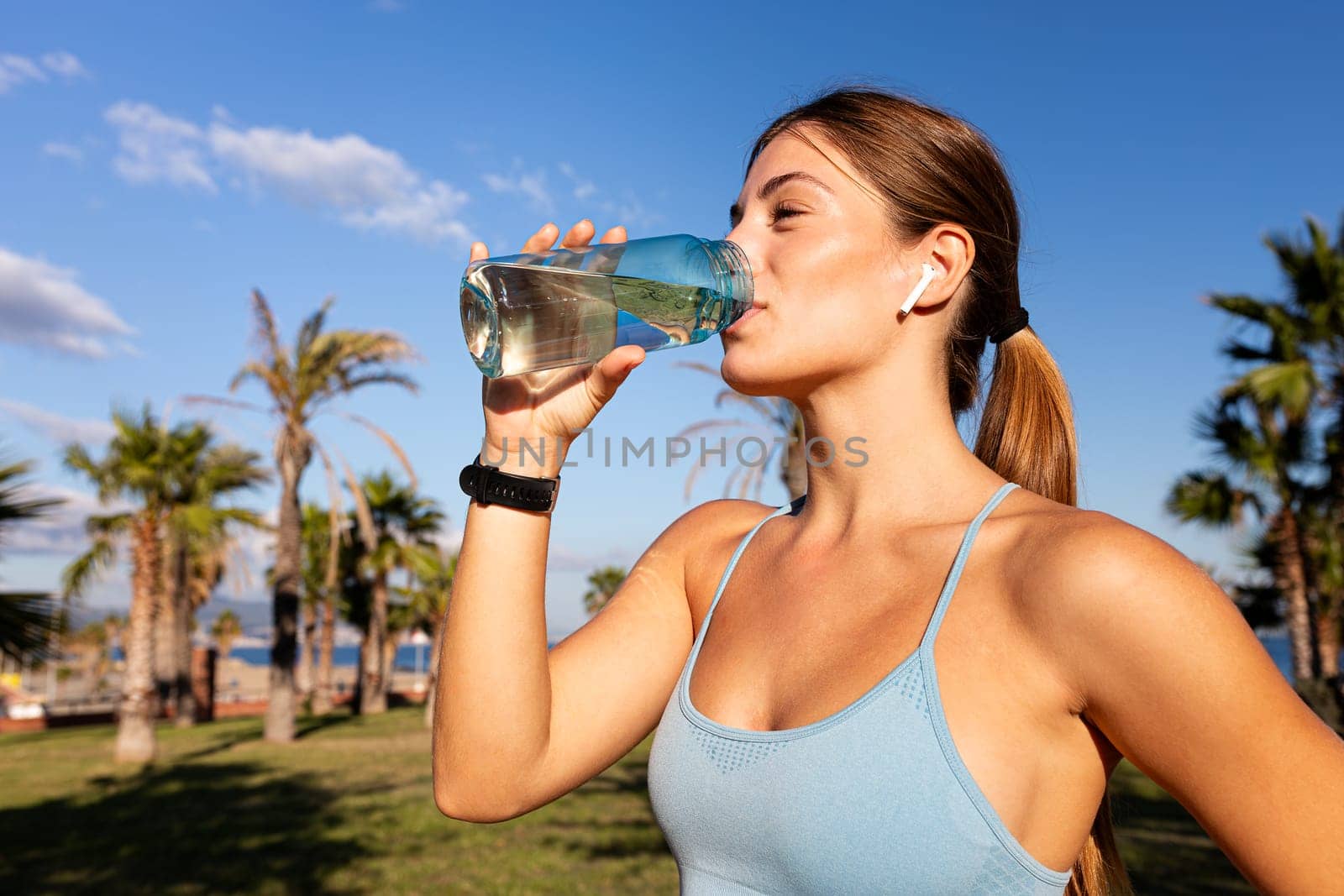 Close up of caucasian female jogger taking a break to hydrate. Young woman drinking water after exercising outdoors. by Hoverstock