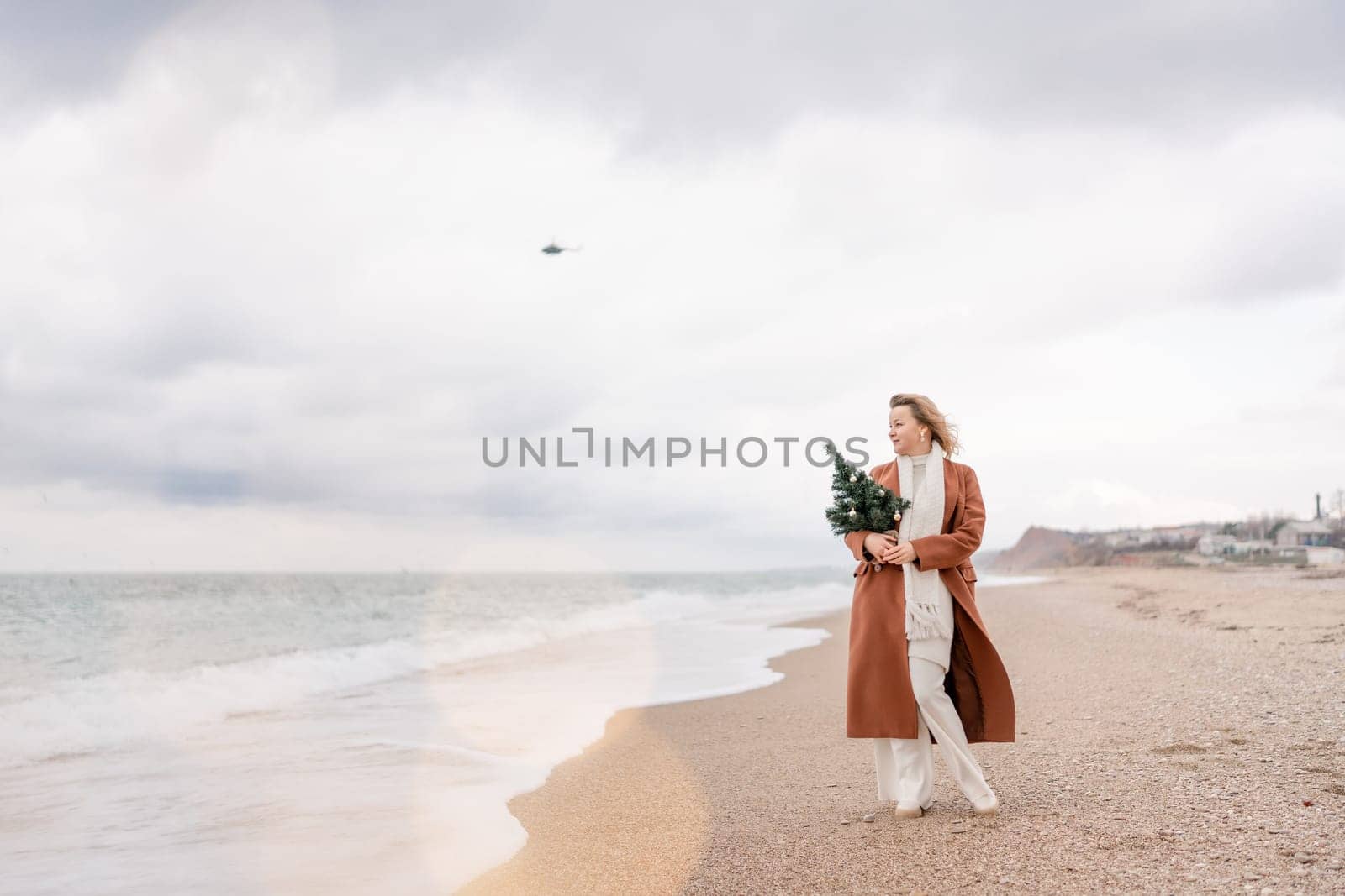 Blond woman Christmas sea. Christmas portrait of a happy woman walking along the beach and holding a Christmas tree in her hands. She is wearing a brown coat and a white suit