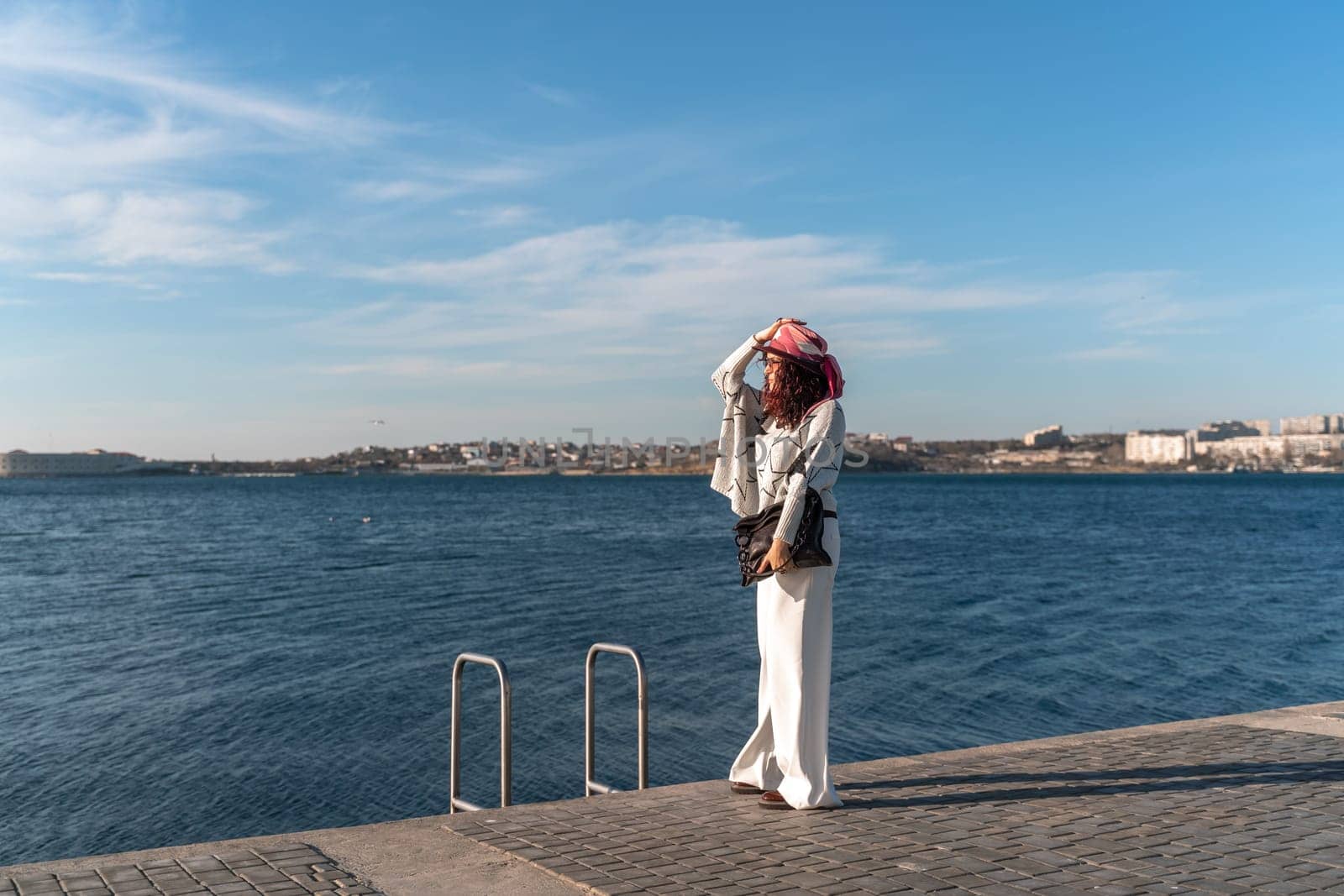 Stylish seashore woman. Fashionable woman in a hat, white trousers and a light sweater with a black pattern on the background of the sea