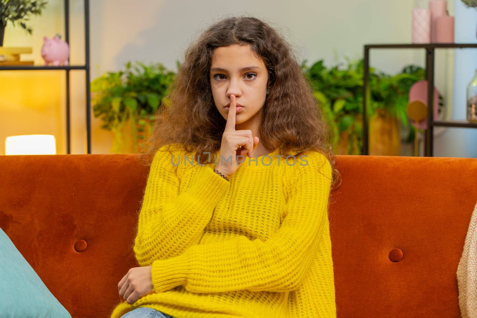 Shh be quiet please. Child girl presses index finger to lips makes silence hush gesture sign do not tell gossip secret. Teenager female 14-15 years kid at home apartment living room sits on couch