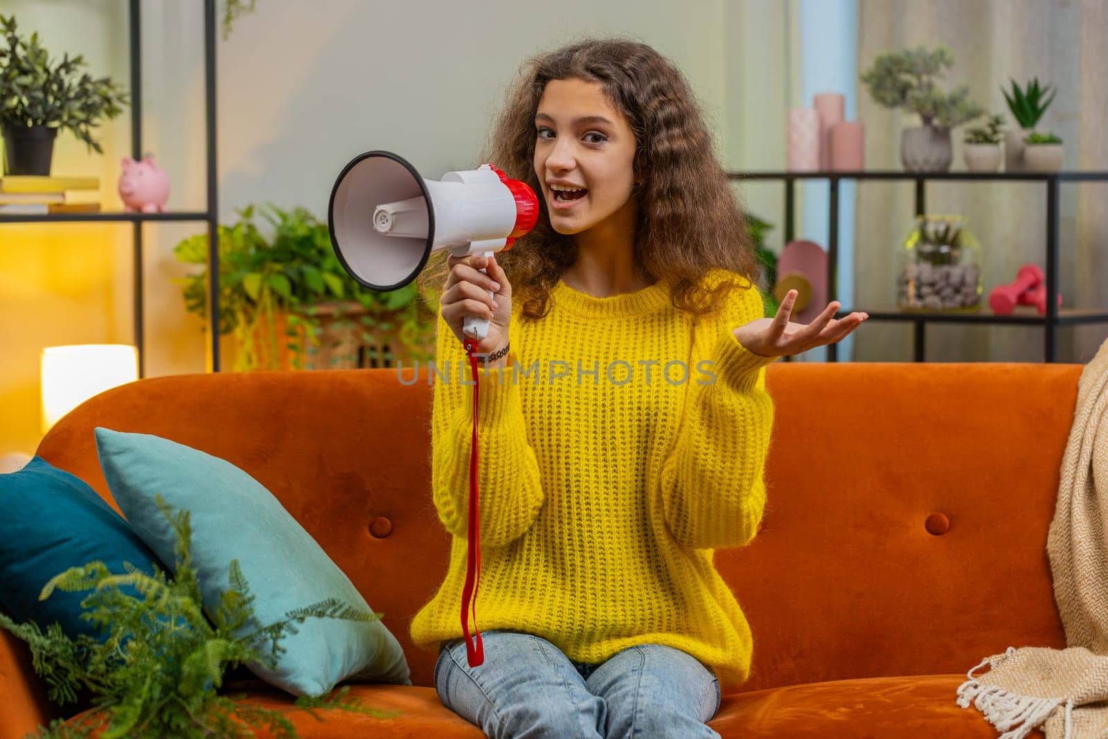 Child girl talking with megaphone, proclaiming news, loudly announcing advertisement warning using loudspeaker to shout speech. Teenager female 14-15 years kid at home living room apartment on sofa