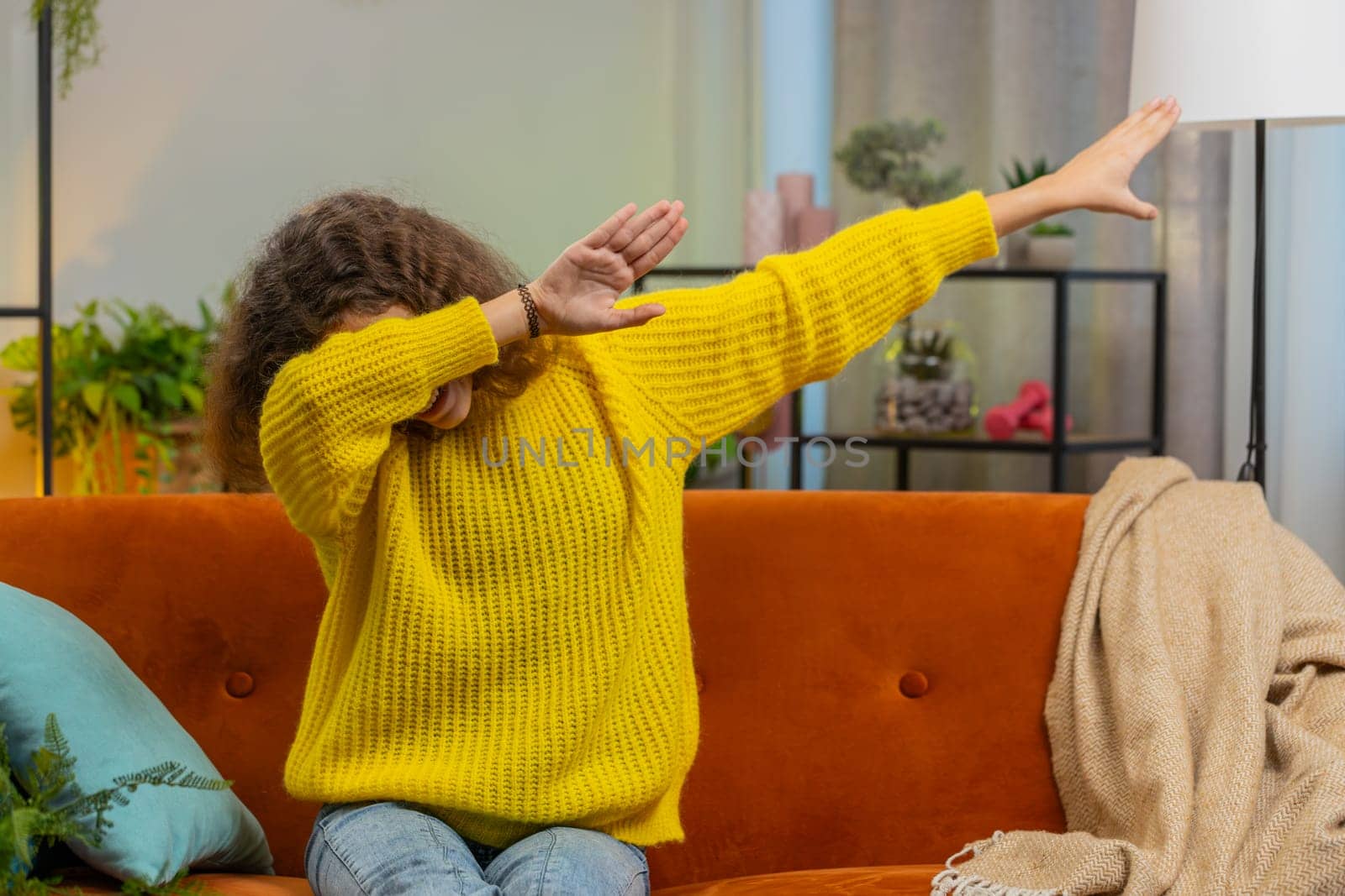 Trendy cheerful positive 14-15 years girl having fun dancing, moving to rhythm dabbing raising hands making dub dance gesture. Teenager Caucasian child kid at home in play living room sitting on couch