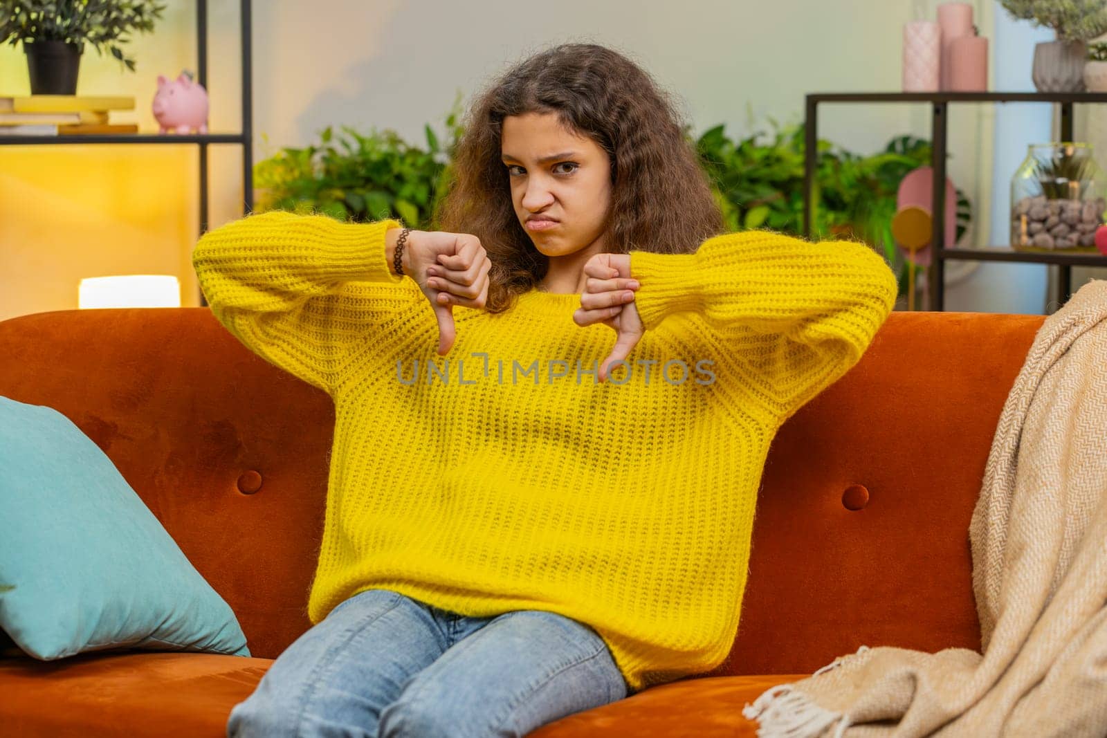 Dislike. Upset child girl showing thumbs down sign gesture, expressing discontent, disapproval, dissatisfied bad work at modern home apartment indoors. Displeased teenager 14-15 years kid on room sofa