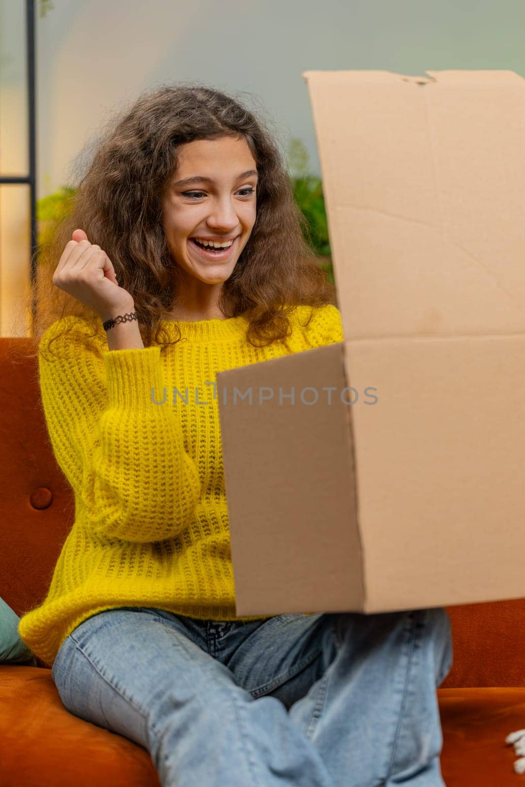 Happy young child girl unpacking delivery parcel. Smiling satisfied teenager kid shopper, online shop customer opening cardboard box receiving purchase gift by fast postal shipping at home. Vertical