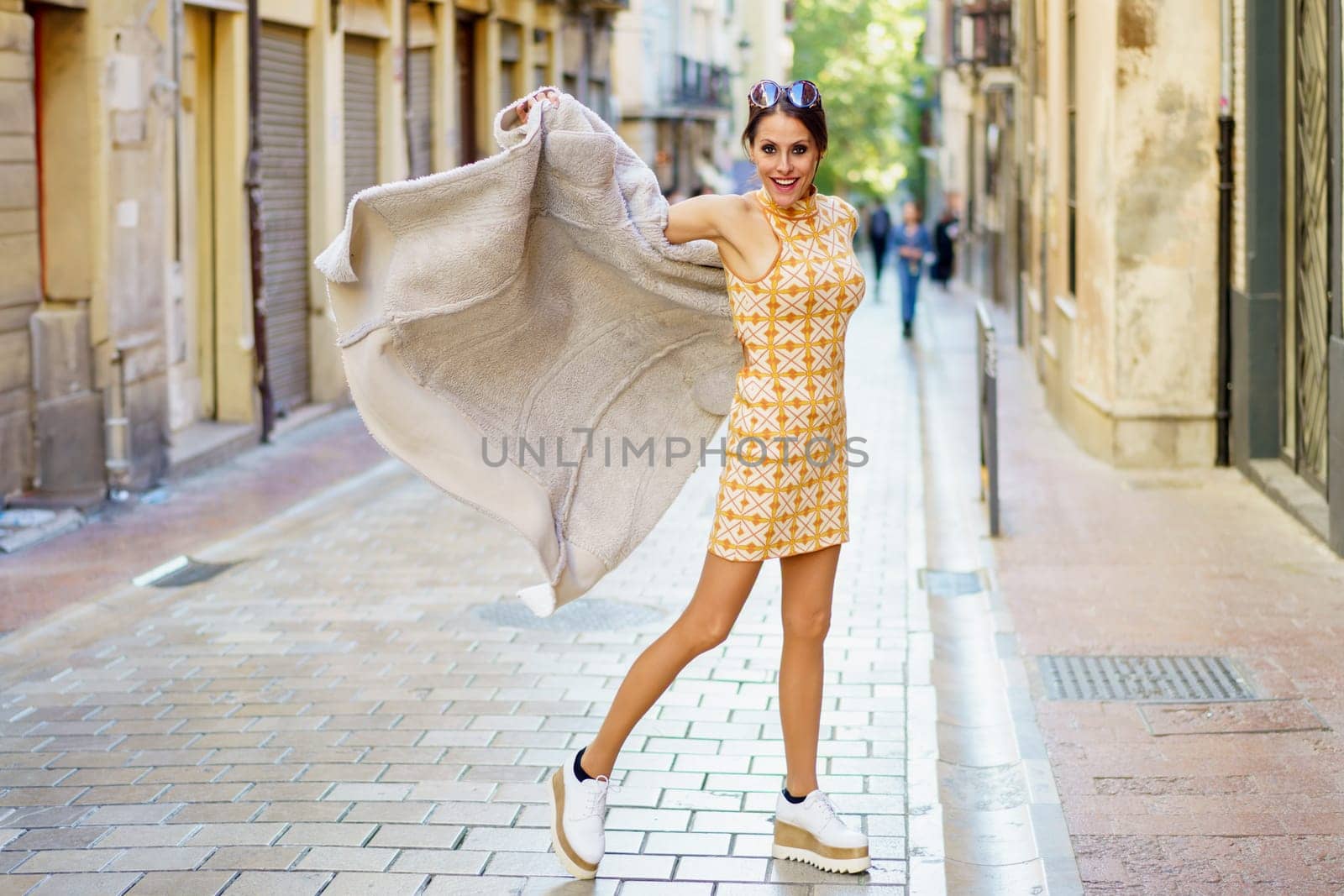 Cheerful young stylish female wearing dress and sneakers with sunglasses standing on paved street against blurred buildings and holding long coat in hands