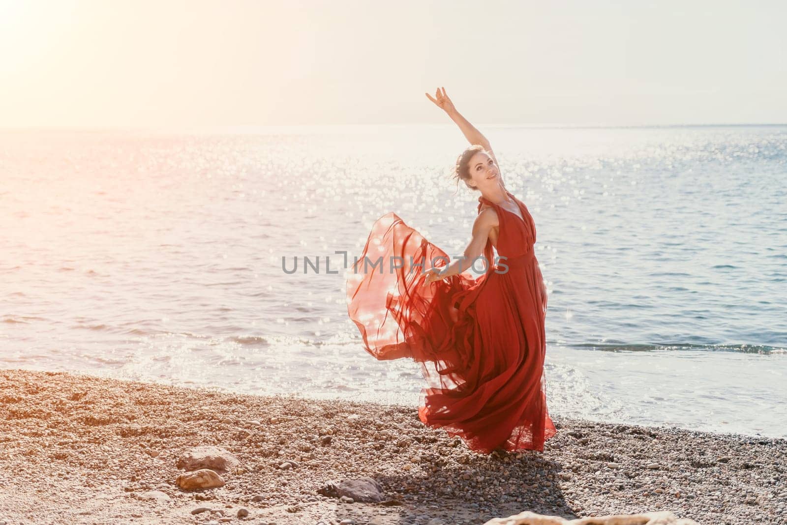 Side view a Young beautiful sensual woman in a red long dress posing on a rock high above the sea during sunrise. Girl on the nature on blue sky background. Fashion photo.