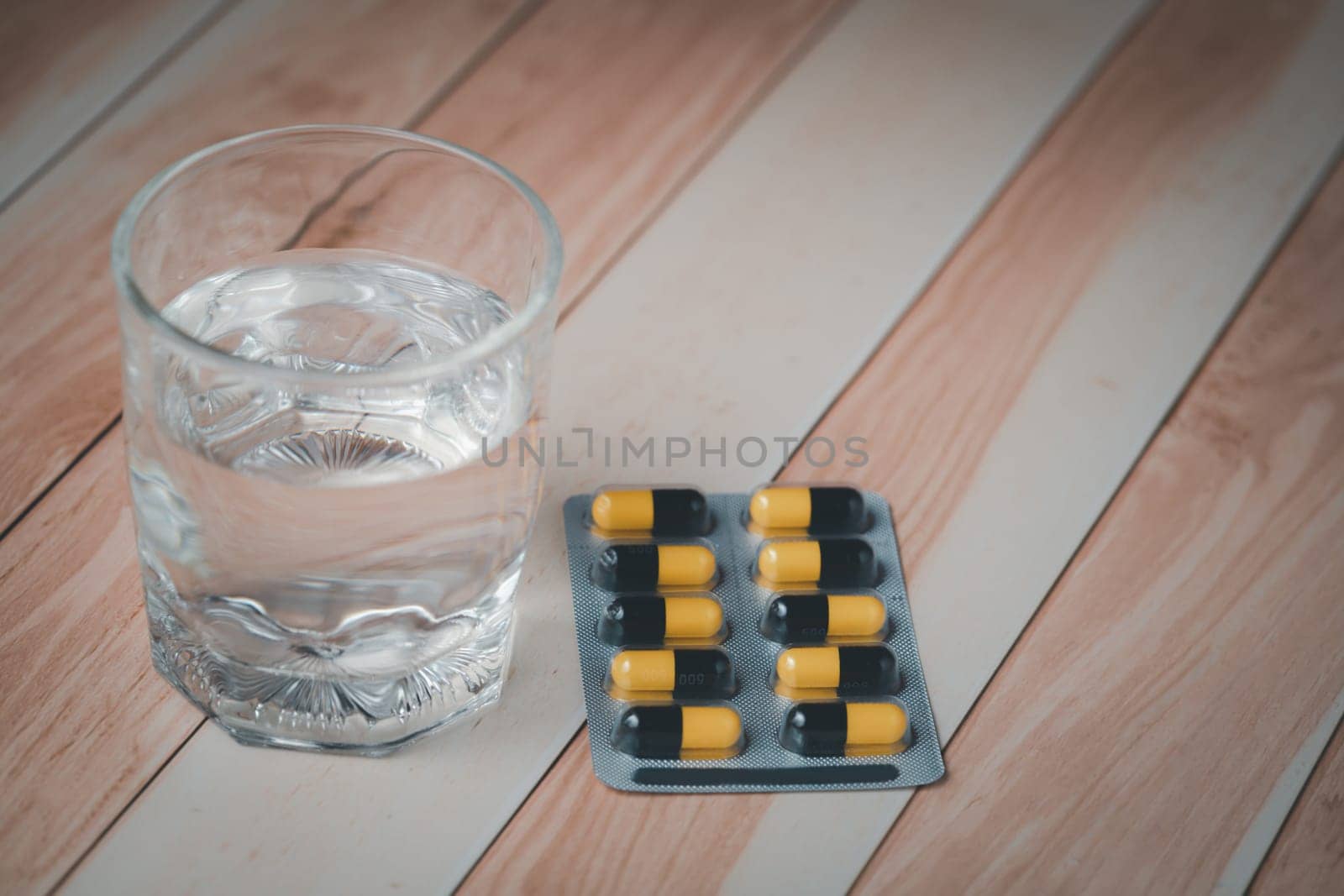 A glass of water and a blister pack of pills, essential for medication, rest on a wooden table, indicating health care routine or treatment
