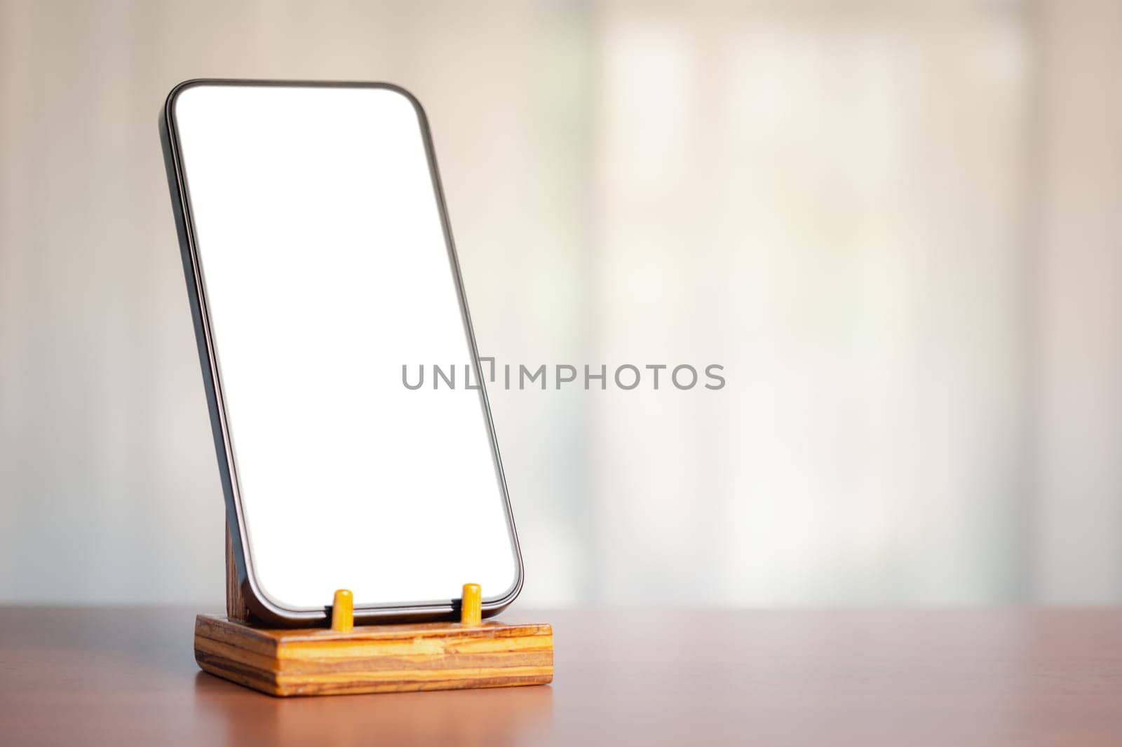 A smartphone with a blank white screen on a wooden stand against a blurred background, bathed in soft sunlight, exemplifies the seamless integration of technology and communication in our daily lives