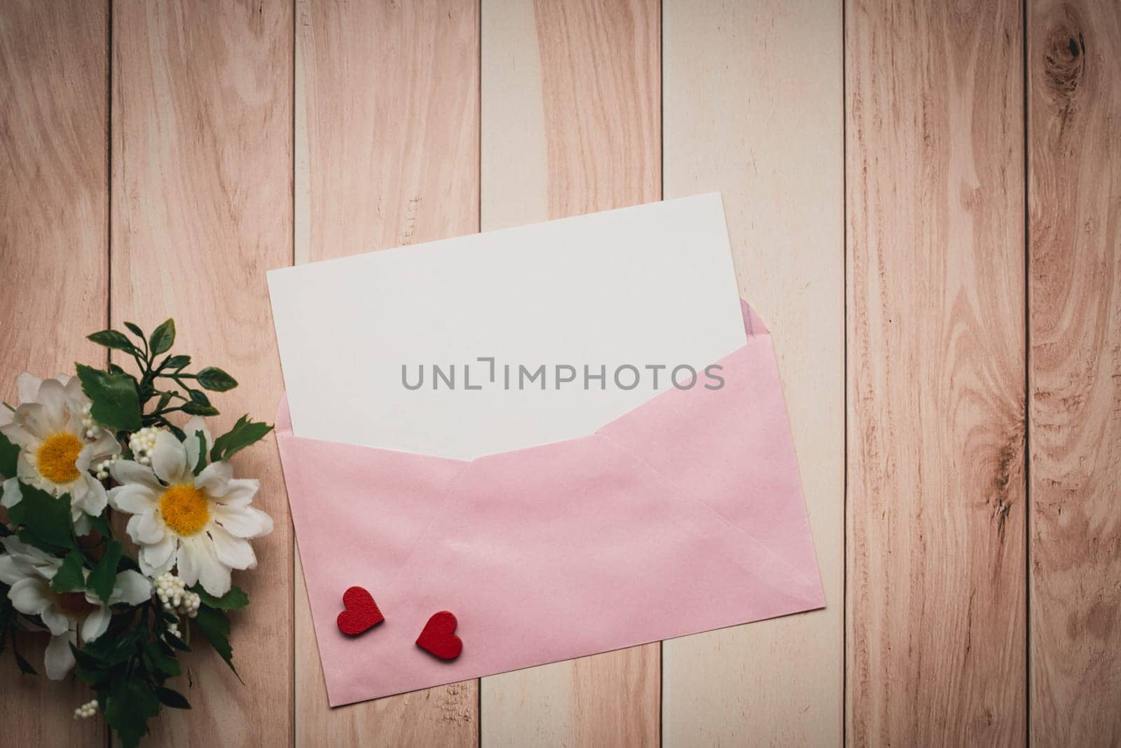 A pink envelope and a white card with a concept of love and Valentine's Day, featuring a red heart and daisy, are gracefully arranged on a wooden table