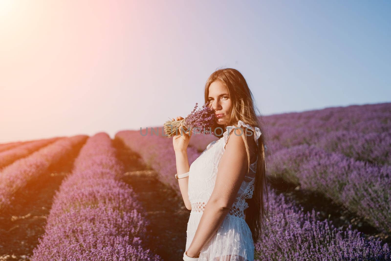 Close up portrait of young beautiful woman in a white dress and a hat is walking in the lavender field and smelling lavender bouquet.