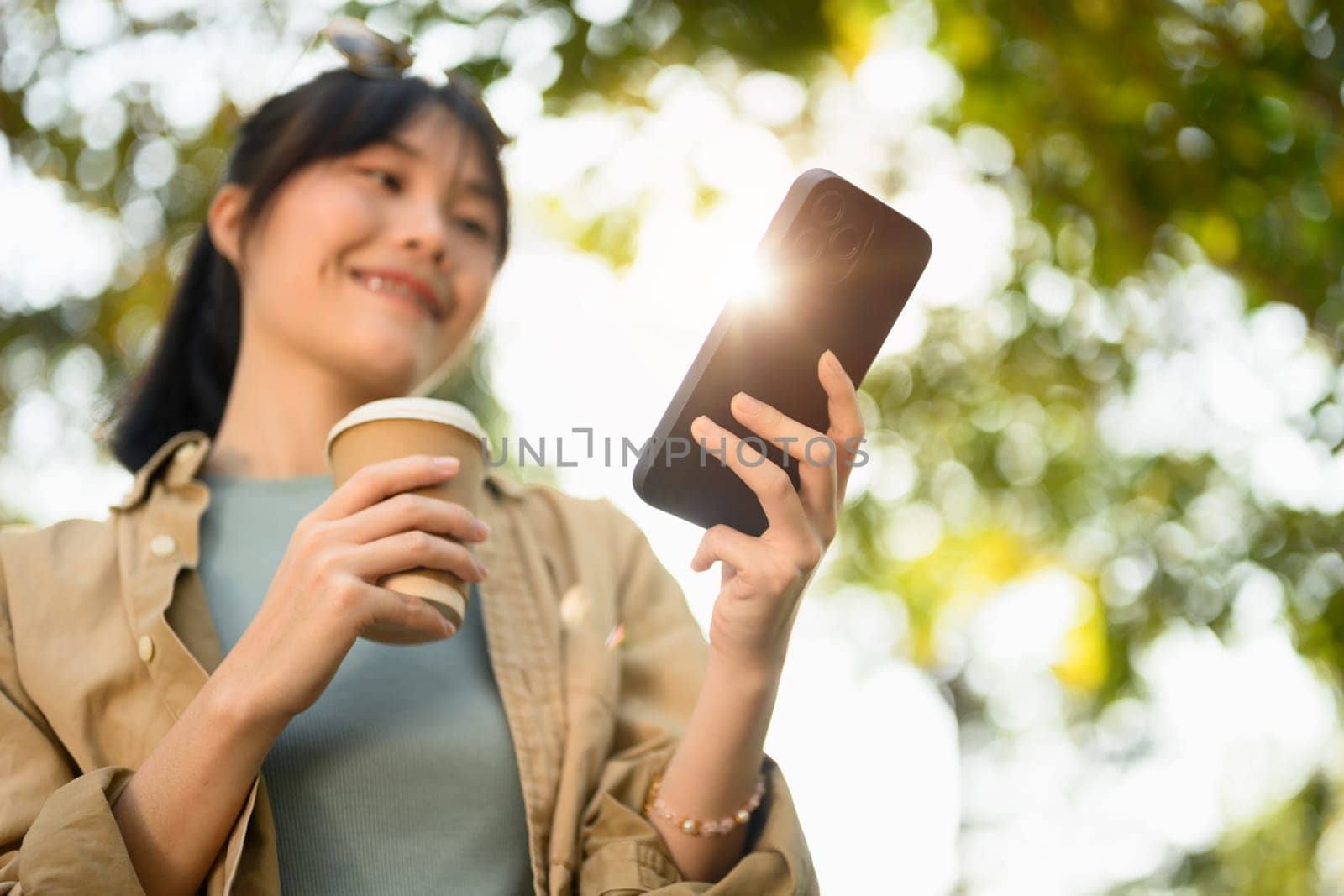 Happy young female freelancer holding paper cup and using smartphone in the summer park by prathanchorruangsak