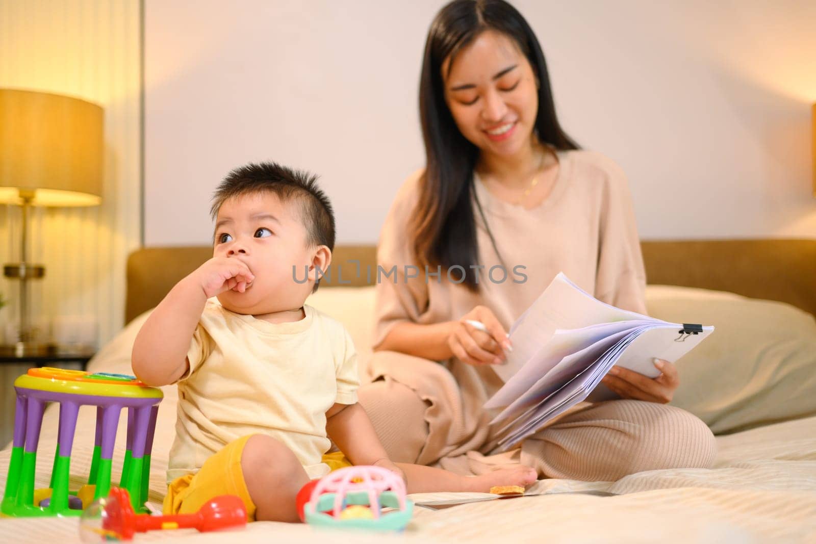 Cute baby boy playing toys on the bed next to her mother working with documents.