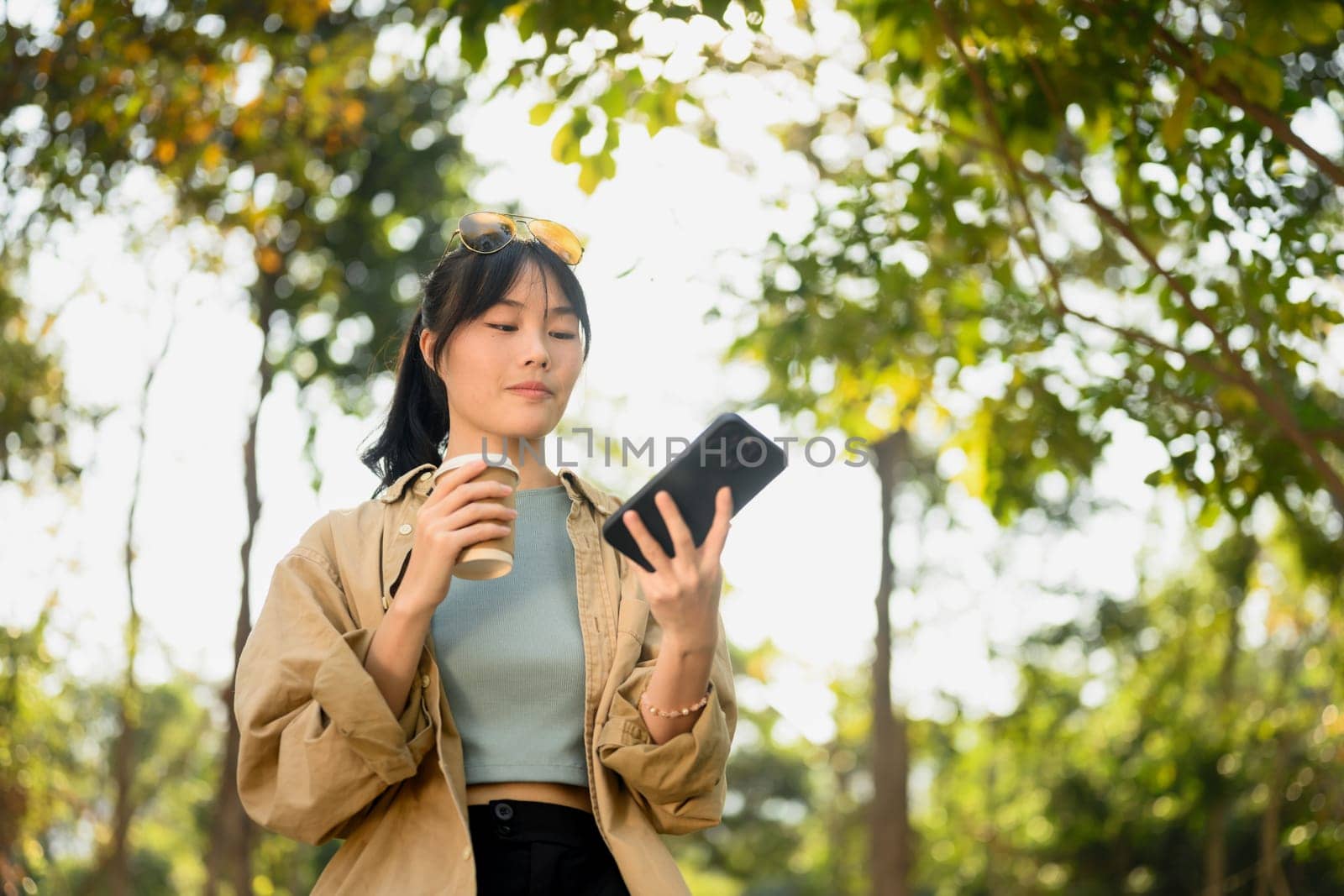 Portrait of beautiful young woman drinking coffee from paper cup and using mobile phone at the park.
