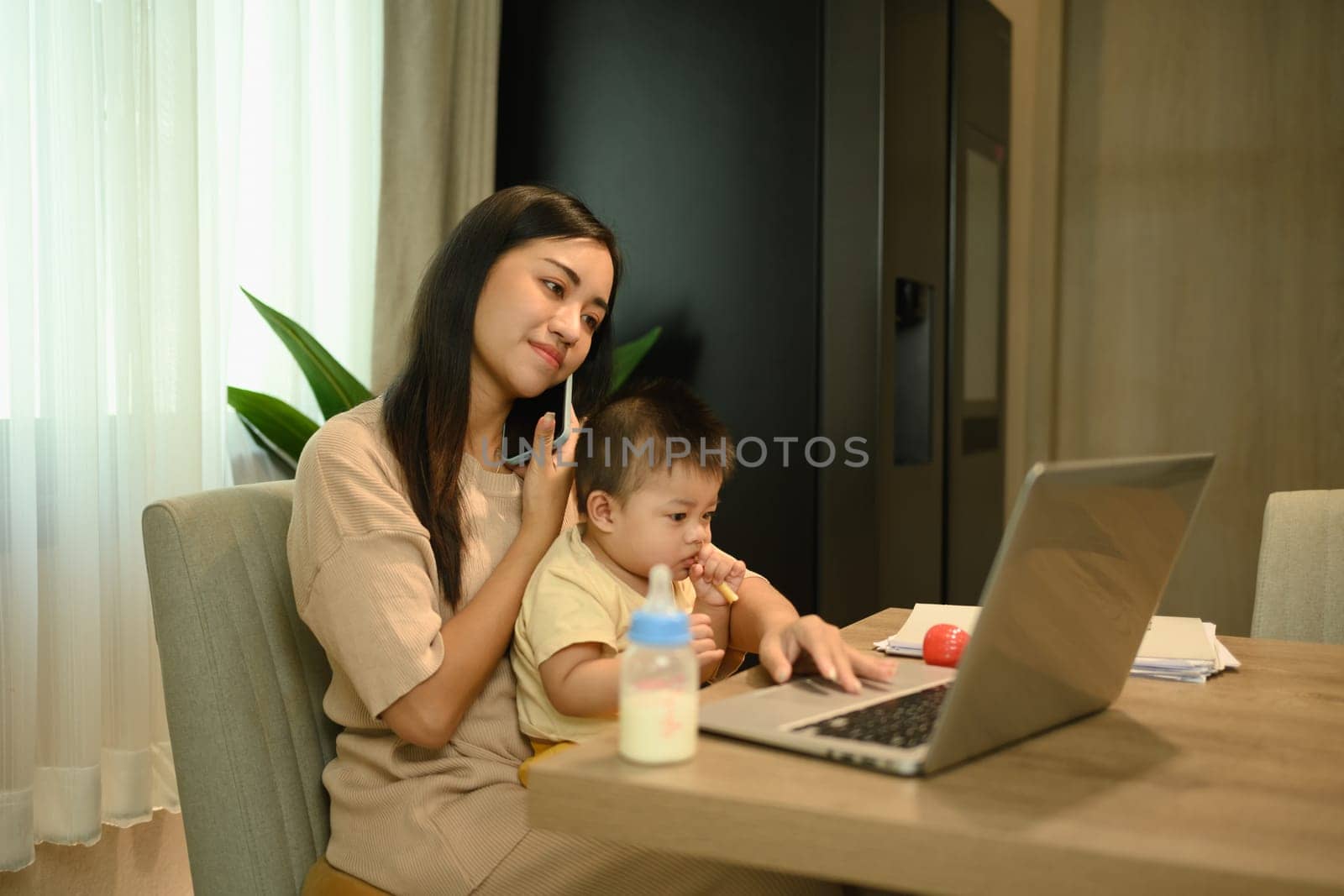 Woman working with laptop and talking on mobile phone while taking care about little baby son by prathanchorruangsak