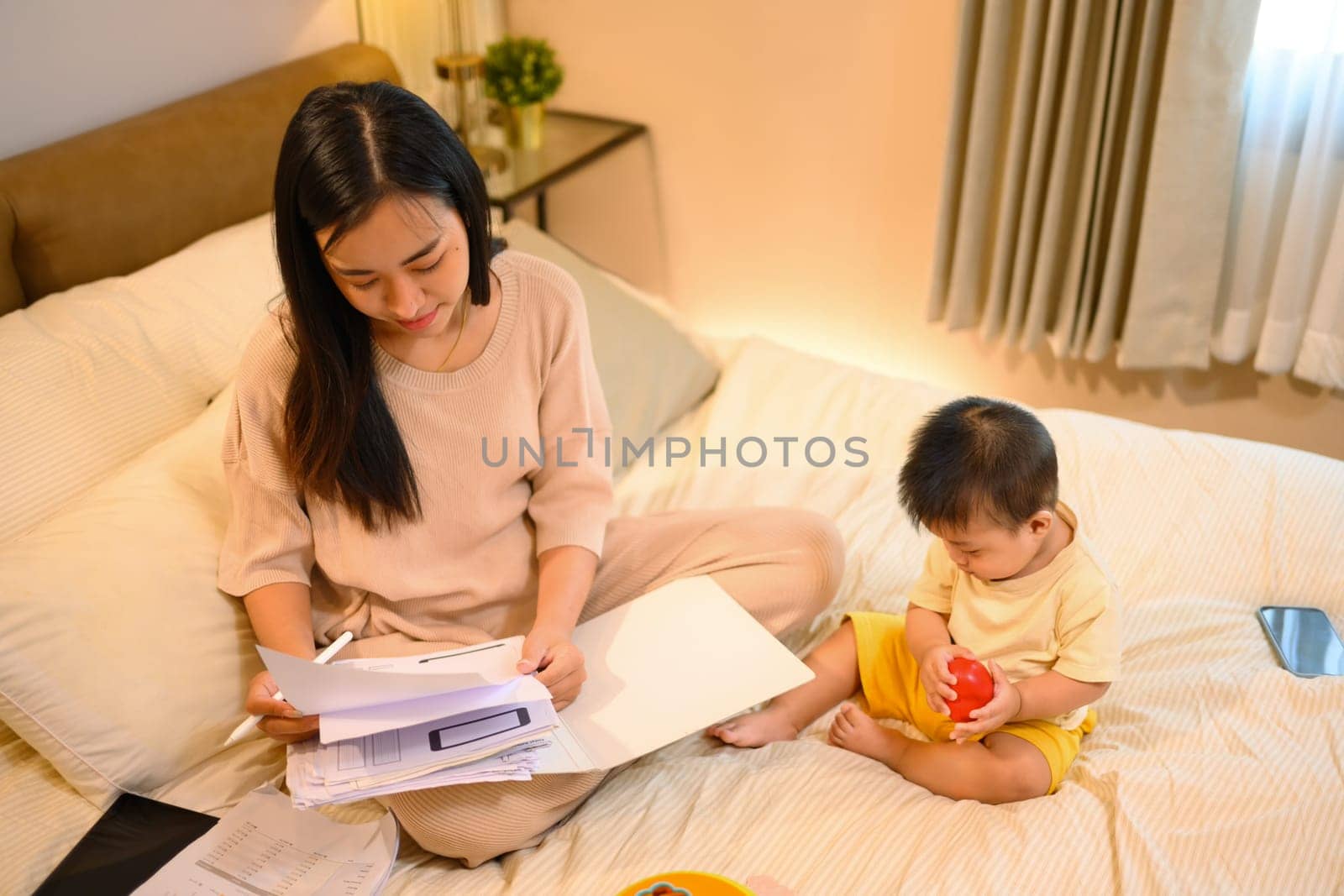 Beautiful business mom working with documents on on bed with her baby boy at home by prathanchorruangsak