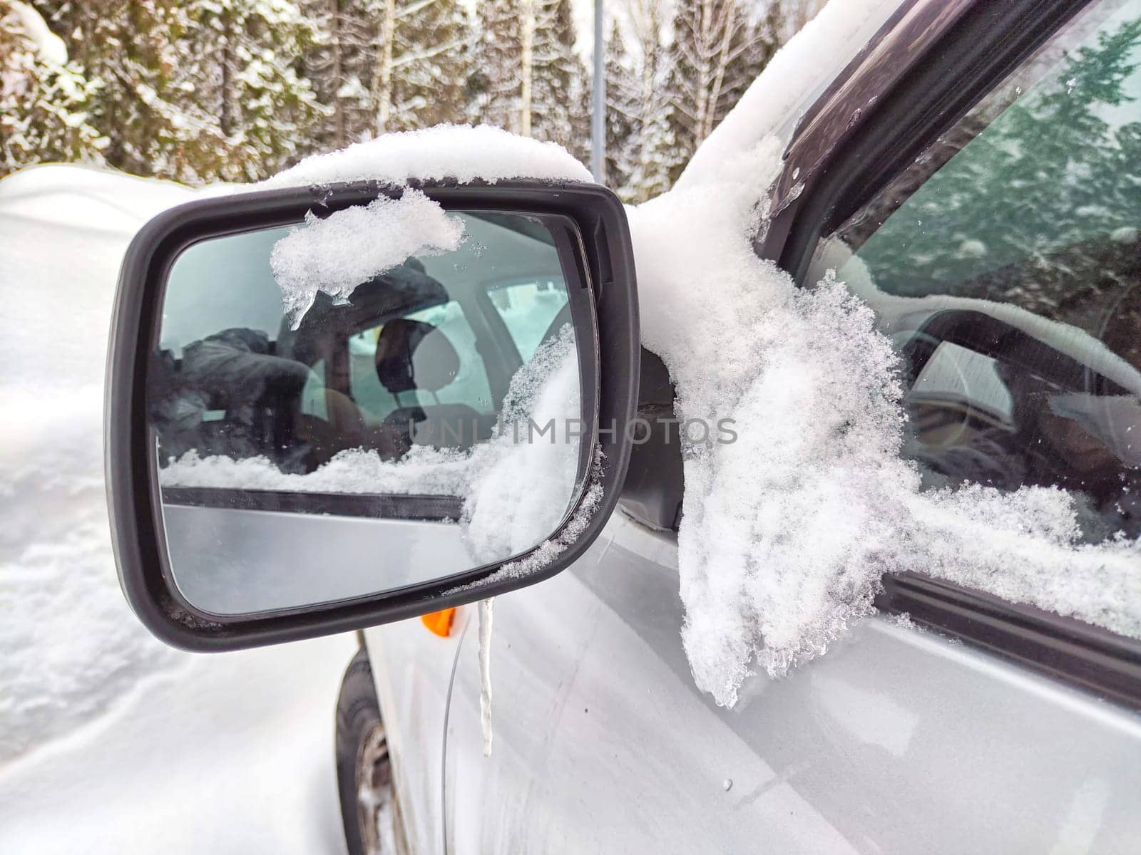 A car parked in a snowy area is seen with its side mirror covered in snow, reflecting the surrounding snow-laden trees and a wintry landscape, highlighting the seasons frosty conditions