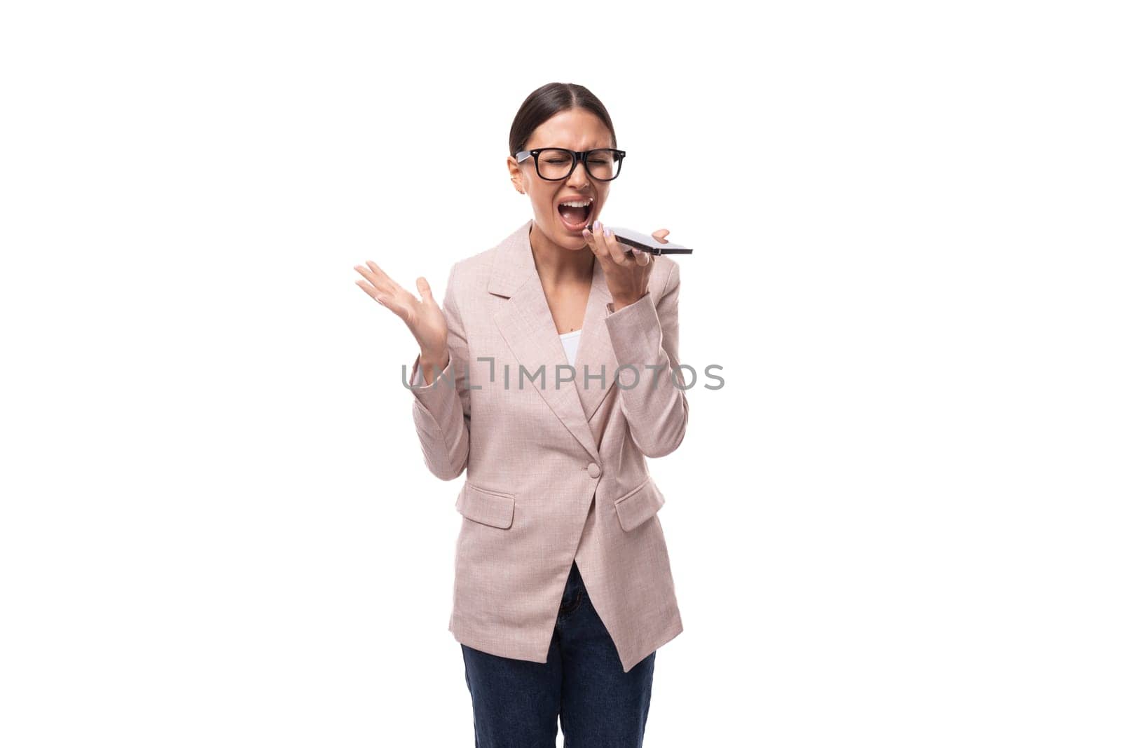 surprised young caucasian woman with gathered black hair in a ponytail dressed in a beige jacket screams into a smartphone.