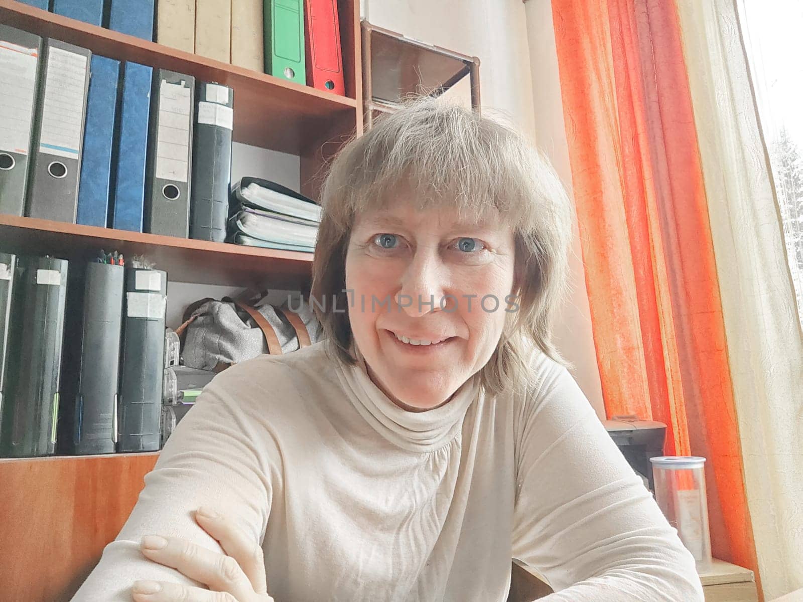 Funny blonde girl or a middle-aged woman poses for the camera with a smile and closet with folders on background. A relaxed Portrait of an Accountant, manager, blogger, freelancer in the workplace