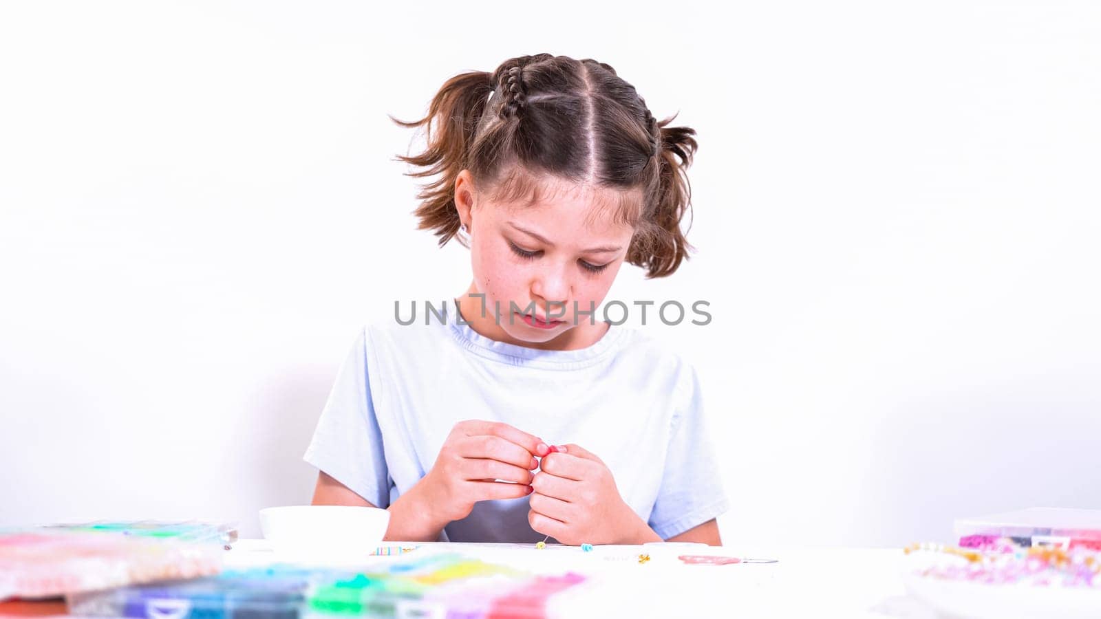 Delicate fingers of a young girl navigate through a treasure trove of bright, multicolored beads, each compartment revealing a new hue to choose from. She's immersed in the joyful task of stringing together a handmade bracelet, with golden beads and pearls lying nearby to add a touch of sparkle to her creation.