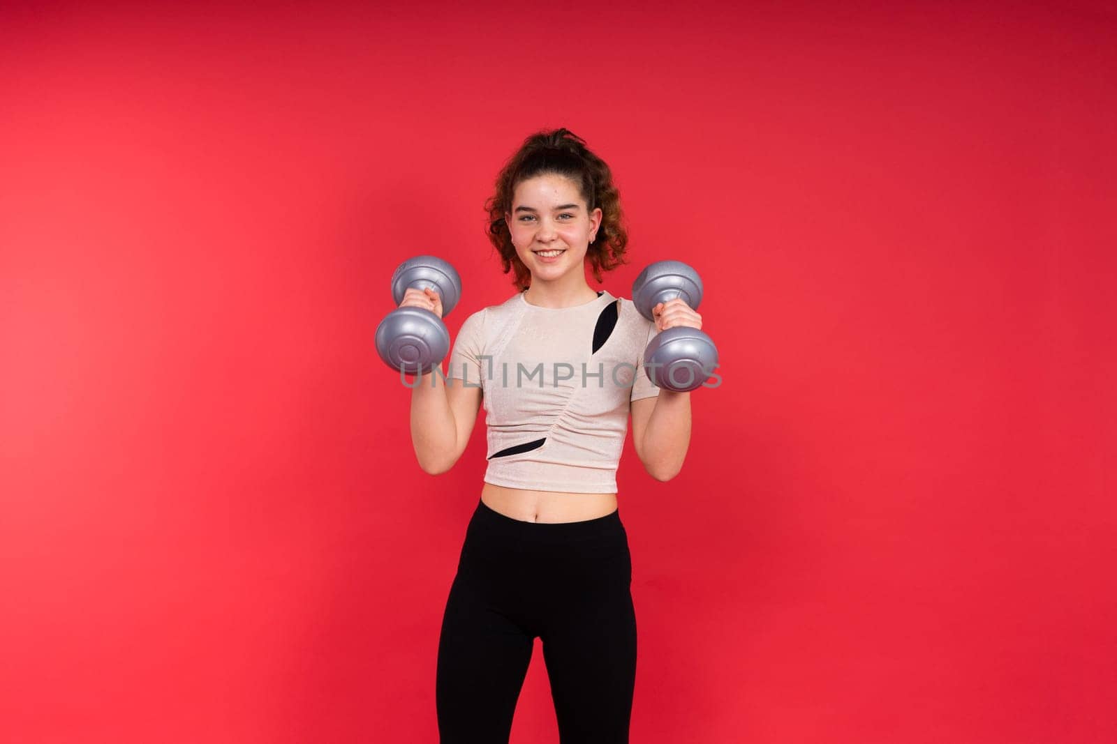 Teenage sportive girl exercises with dumbbells to develop muscles isolated on a red background.