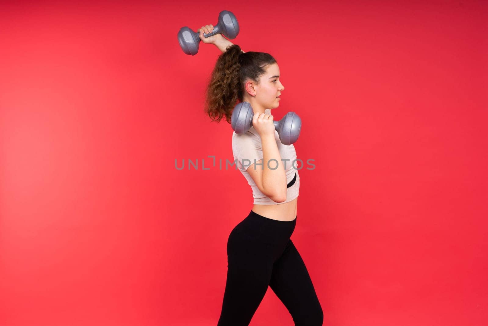 Teenage sportive girl exercises with dumbbells to develop muscles isolated on a red background.