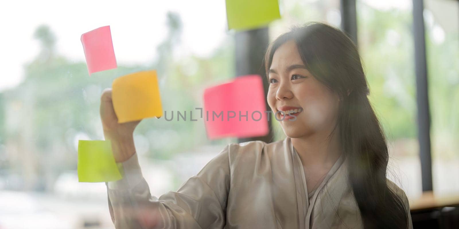 Smiling businesswoman Asian people thinking and use sticky notes on glass wall in office by nateemee