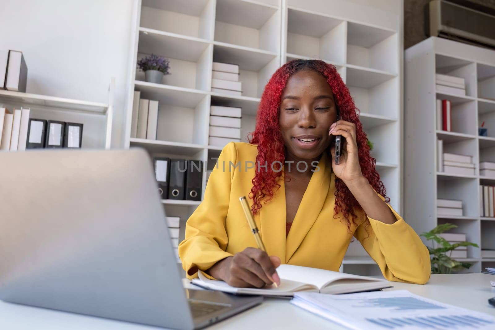 Young African American business woman talking on the phone and taking notes while working in the office by wichayada