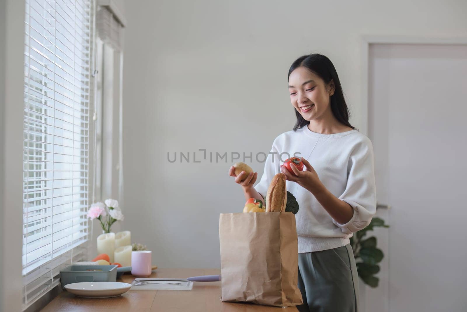 A beautiful young woman unwraps a paper bag with vegetables and fruits and prepares to make a healthy meal in the kitchen. by wichayada
