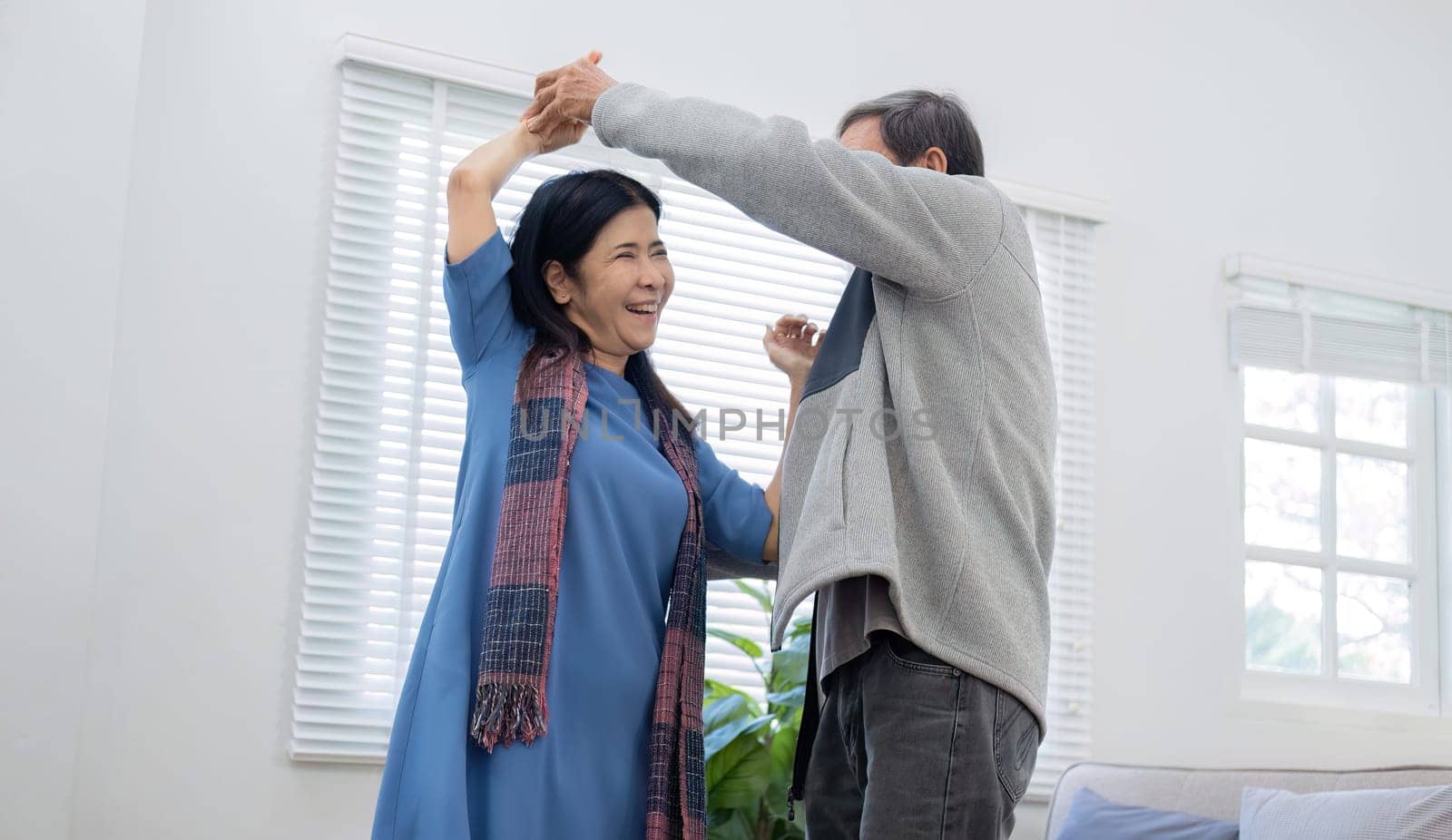 Elderly Asian couple in their 60s dancing in the living room together, enjoying music and smiling in love..