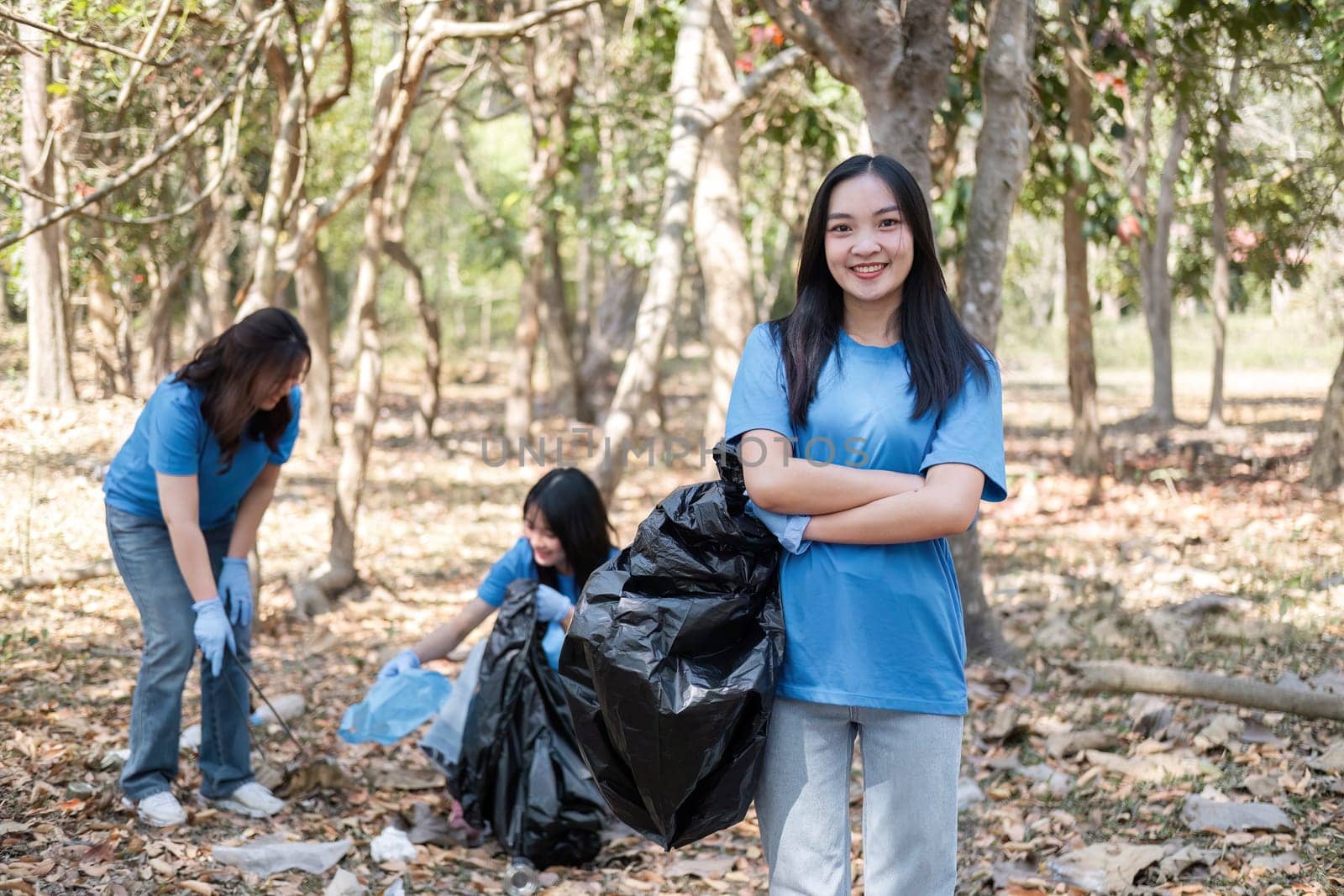 Portrait of a cute young woman holding a garbage bag with a group of Asian volunteers helping to collect garbage in plastic bags, cleaning up the area in the forest to preserve the natural ecosystem..