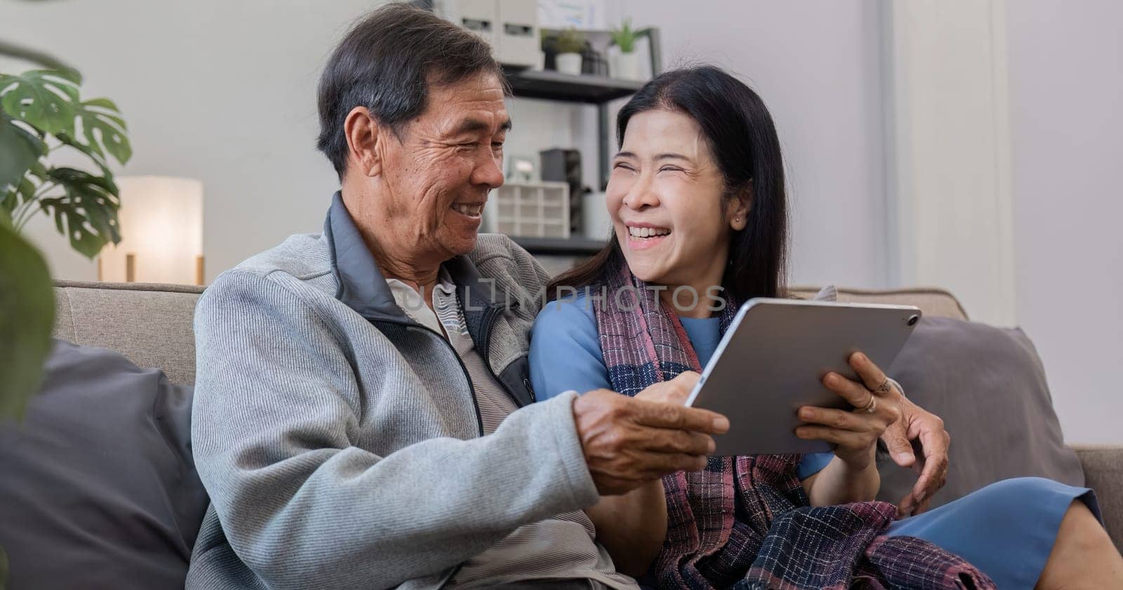 A senior couple in their 60s spends their free time sitting on a tablet, relaxing and watching entertainment programs, relaxing happily together on the sofa in the living room..