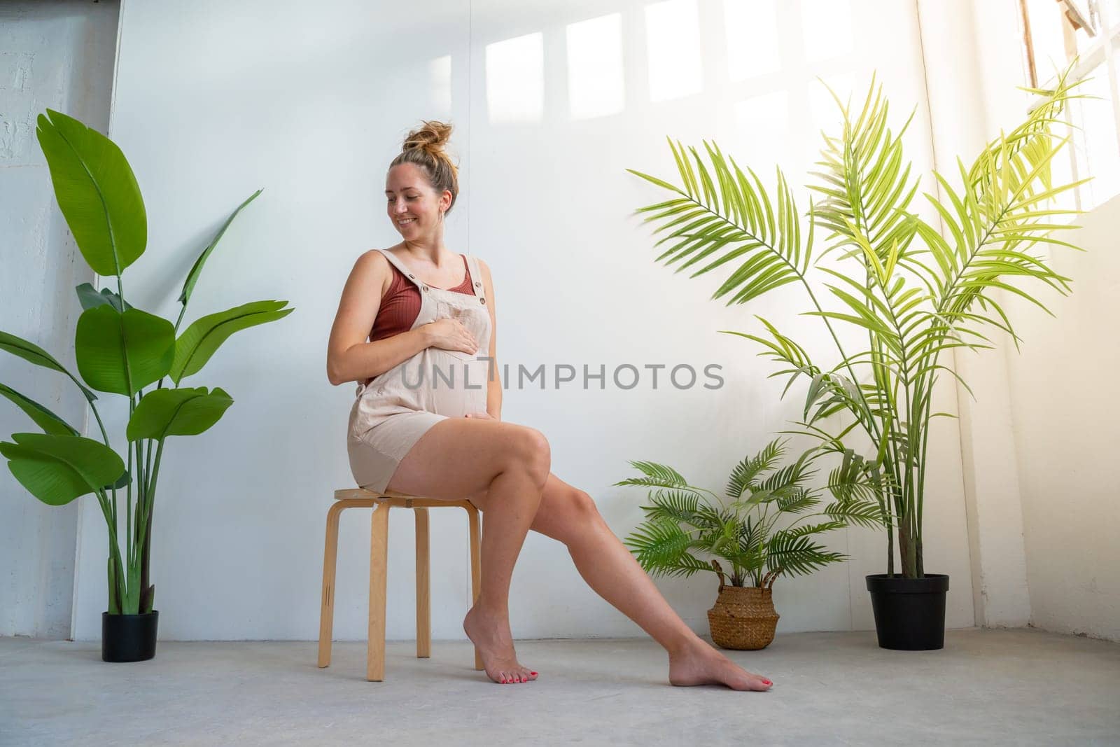 Happy pregnant women touching her belly relaxed on a chair between indoor plants. by PaulCarr