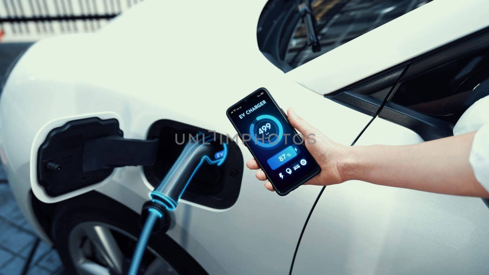 Businessman holding smartphone display battery status interface by smart EV mobile application while EV car recharging electricity from charging station in car park. Peruse