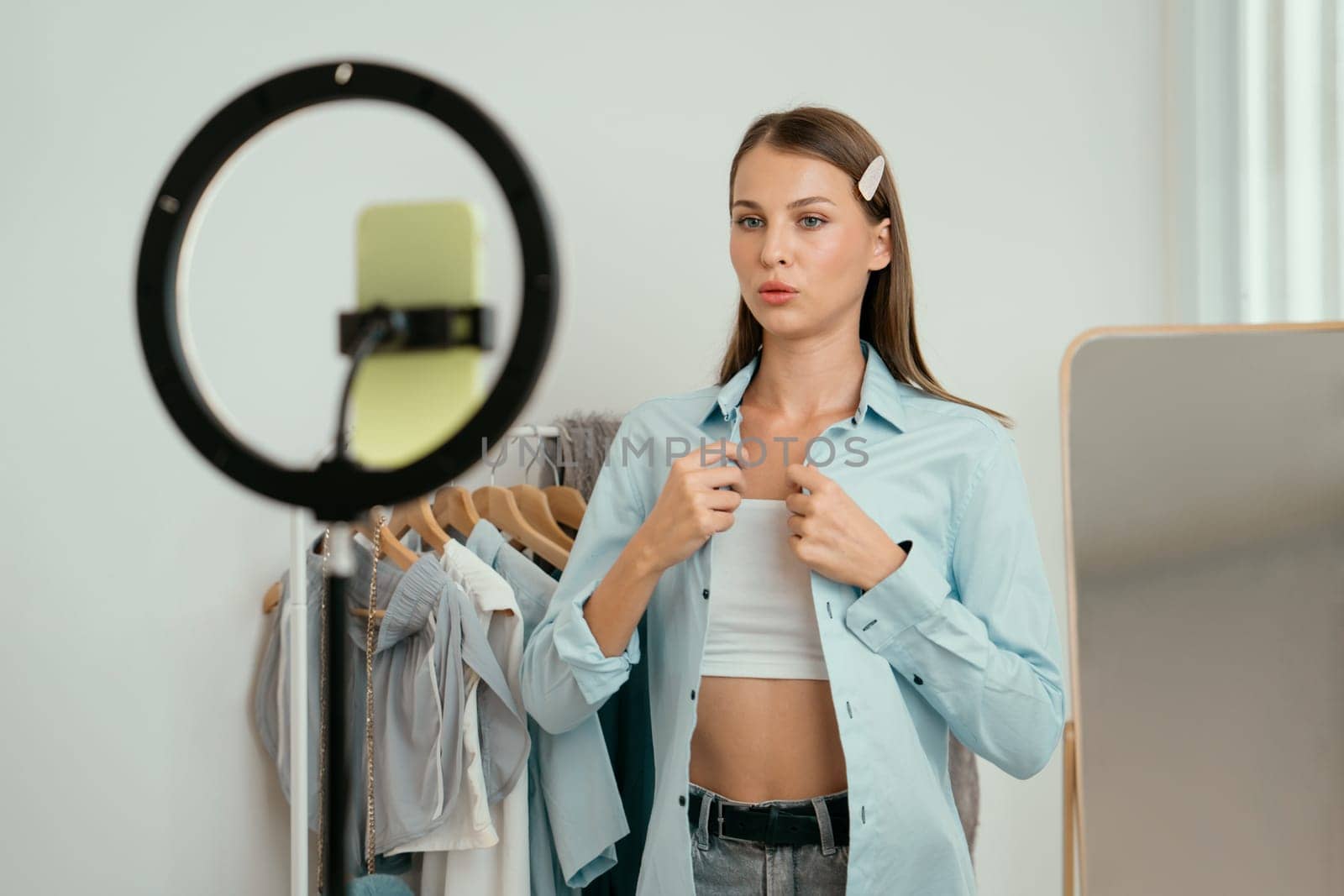 Young woman making fashion video content for social media. Blogger smiles to camera using selfie stick and light ring while making persuasive online clothing sell to audience or followers. Unveiling