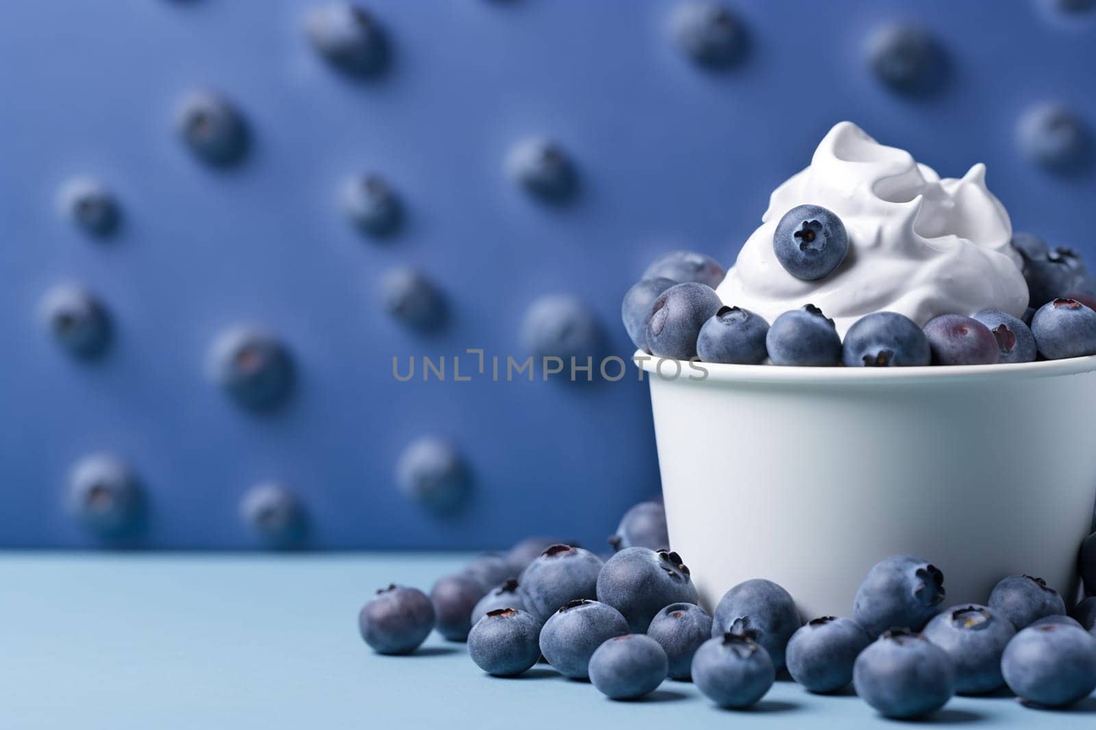 A bowl of whipped cream topped with blueberries on a blue backdrop with scattered berries.