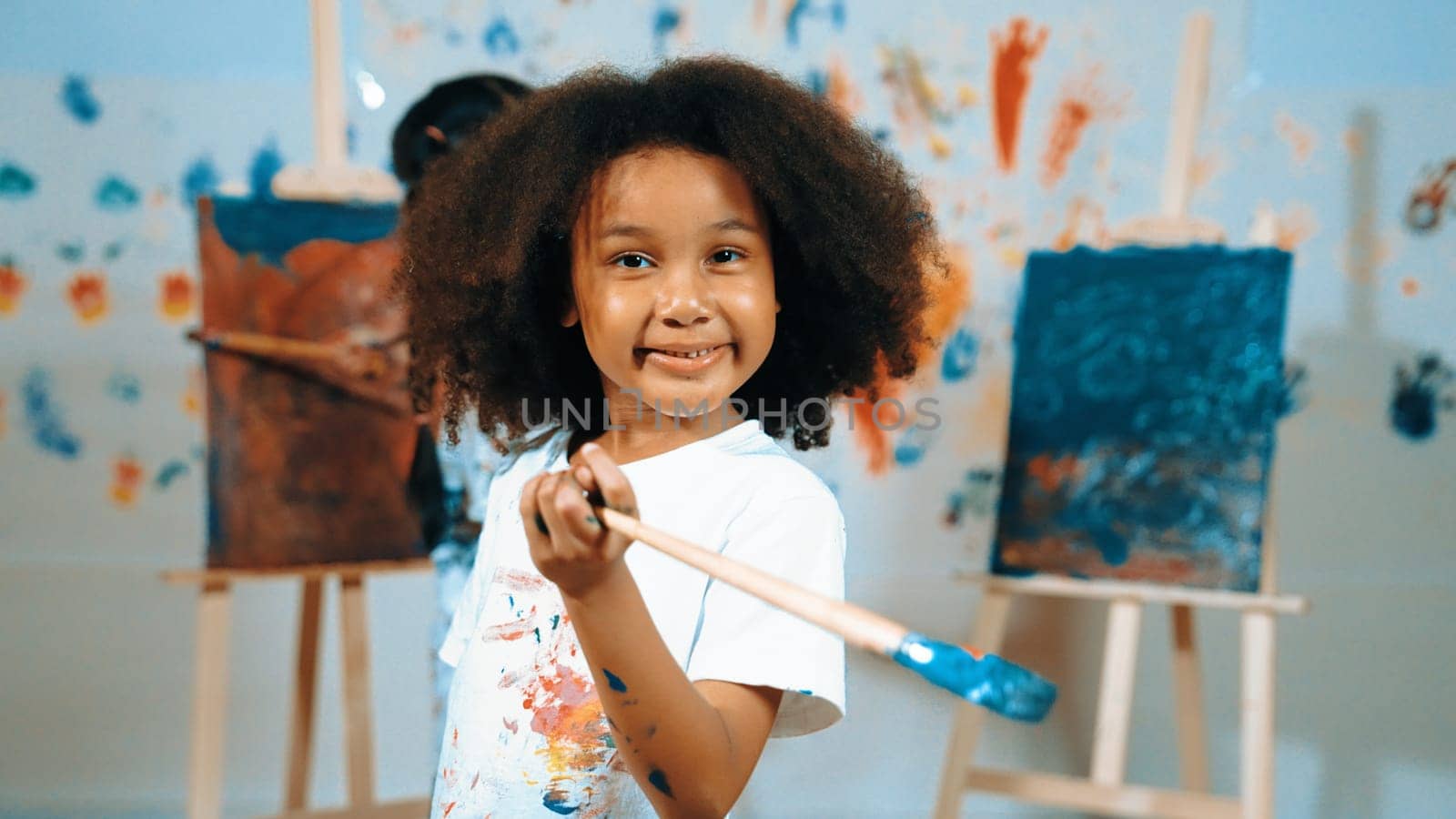 Cute african girl holding painted brush while student drawing canvas behind. Multicultural children attend in art lesson while pretty learner looking at camera. Creative activity concept. Erudition.