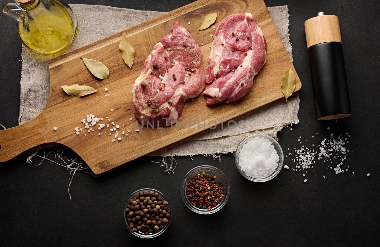 Two raw pork neck steaks on a board and spices for cooking. Top view of black table. Copy space