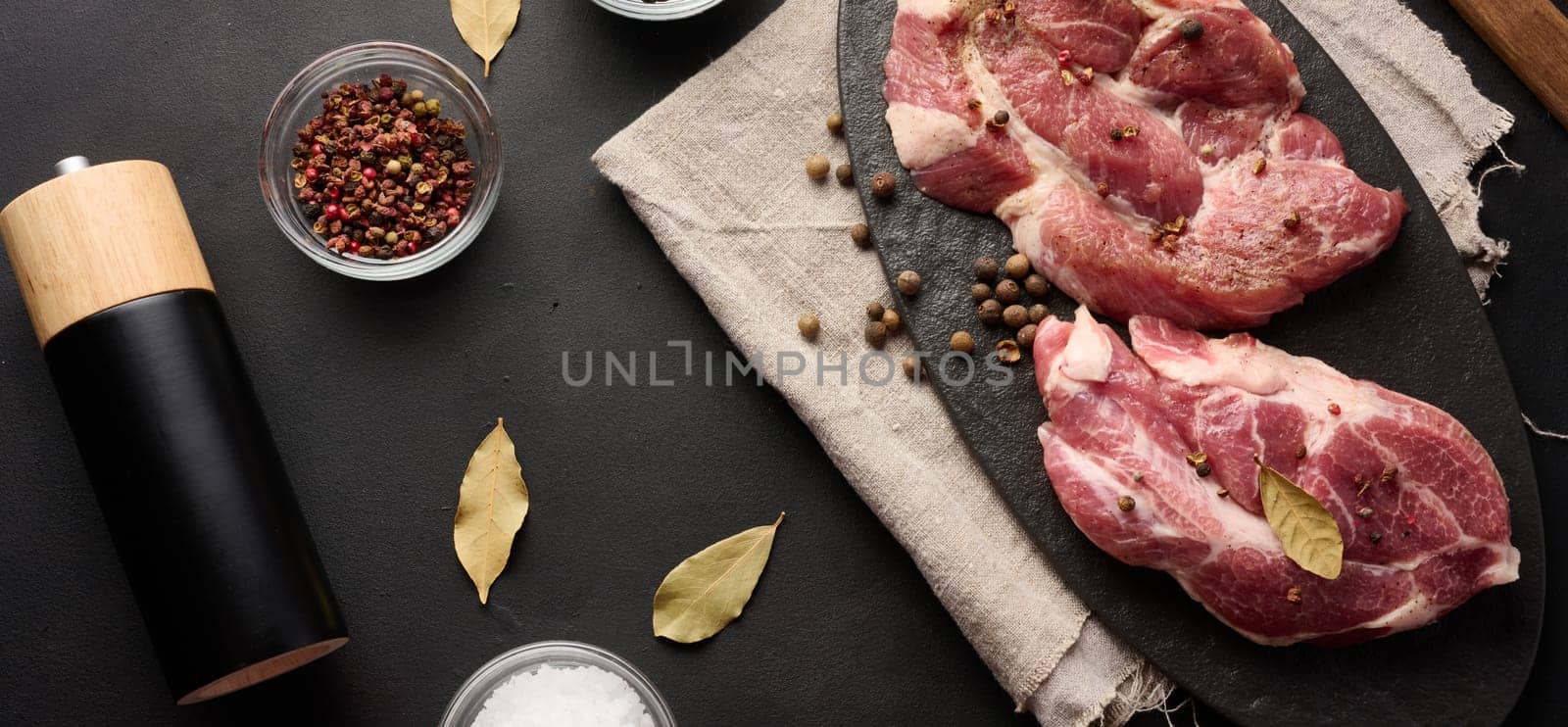 Two raw pork neck steaks on a board and spices for cooking. Top view of black table